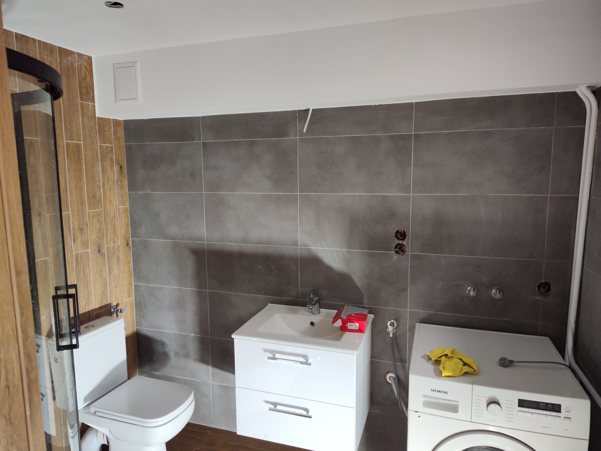 Nowoczesna łazienka z szarą płytką, drewnopodobną kabiną prysznicową, białą szafką z umywalką i pralką Siemens z żółtą ścierką na górze, widoczne niedokończone instalacje na ścianie.