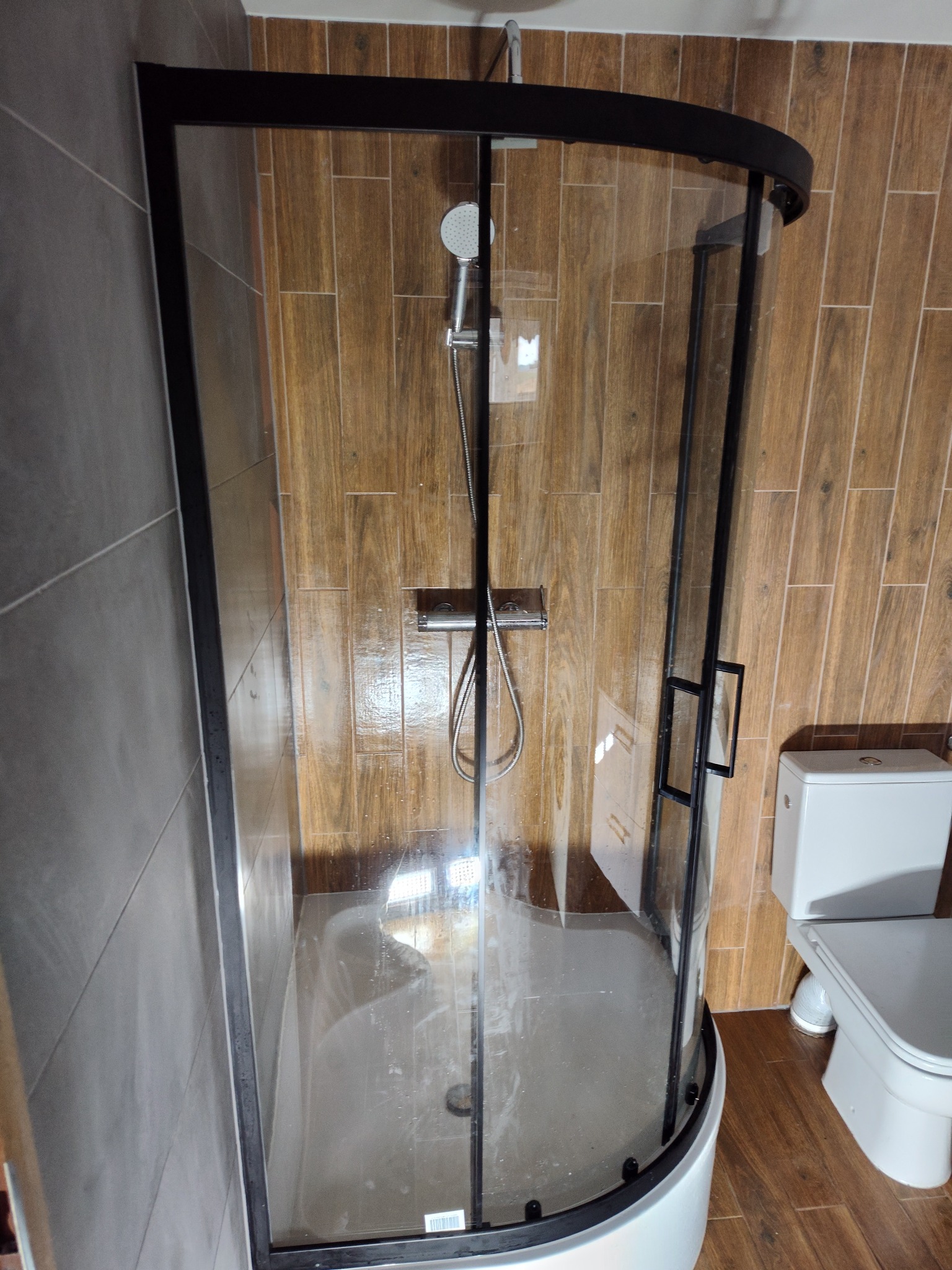 Kabina prysznicowa z czarną ramą na tle drewnopodobnych płytek i szarej ściany, obok toaleta.