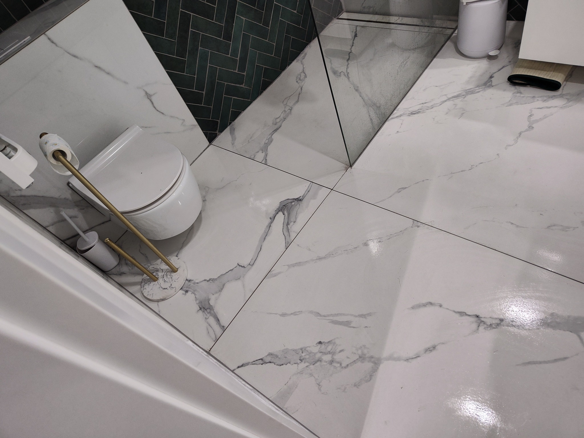 Nowoczesna łazienka z białymi marmurowymi płytkami podłogowymi i ścianą w ciemnozieloną jodełkę, widoczny biały sedes z złotym uchwytem na papier toaletowy i szczotką, oraz fragment białej wanny.