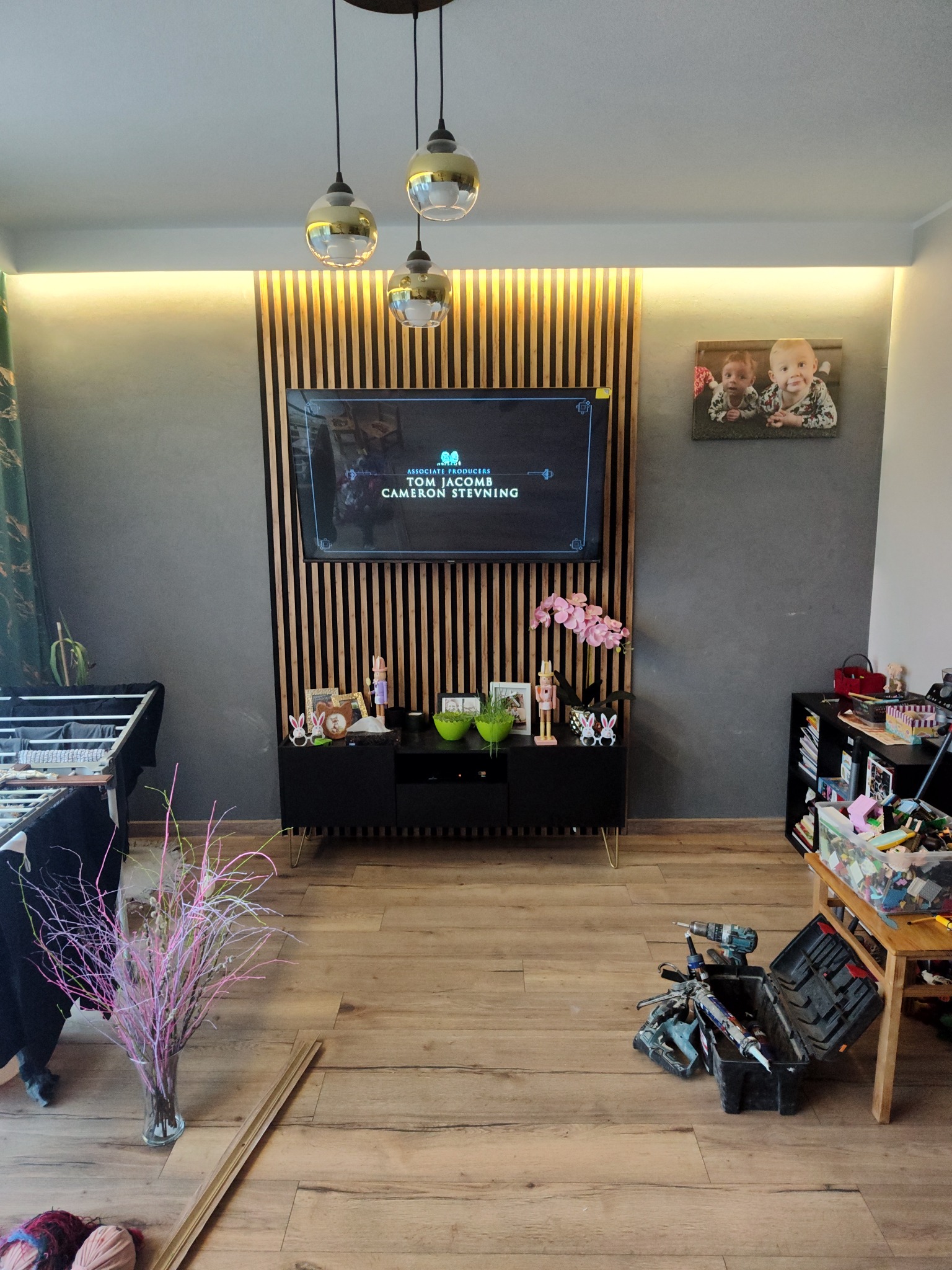 Salon z lamelami za telewizorem, oświetleniem LED i narzędziami na podłodze. Widoczne elementy dekoracyjne i zabawki, sugerujące remont lub wykończenie wnętrza.