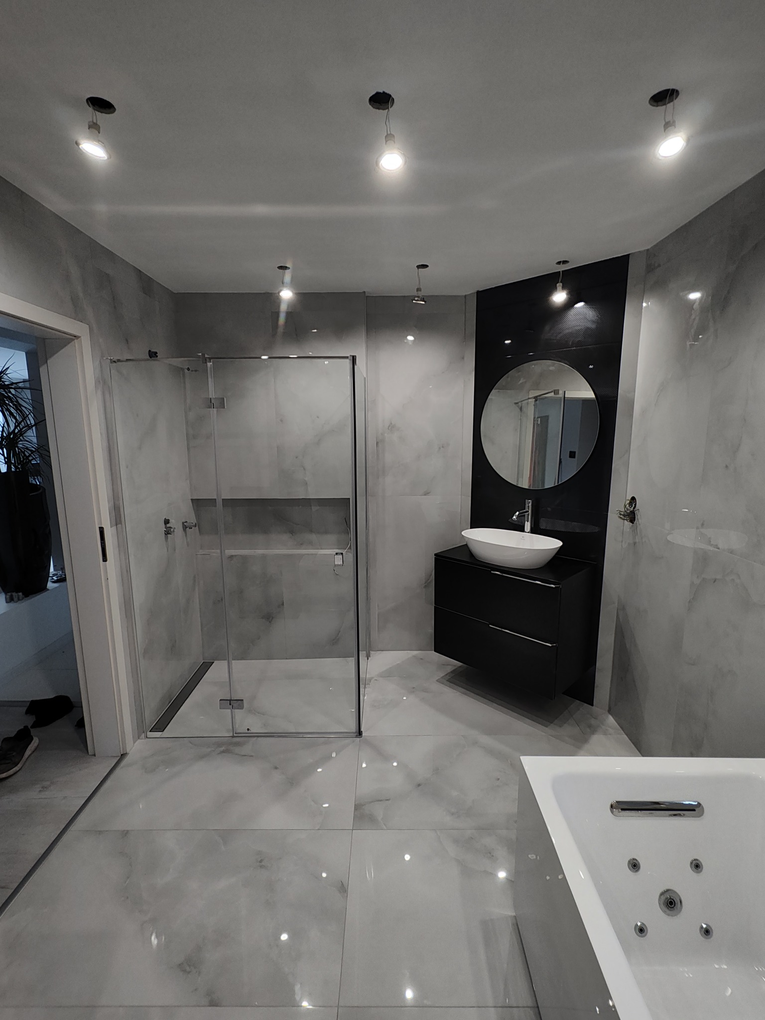 Nowoczesna łazienka w szarościach: kabina prysznicowa ze szklanymi drzwiami, umywalka nablatowa na czarnej szafce, duże lustro w okrągłej ramie, oświetlenie punktowe.