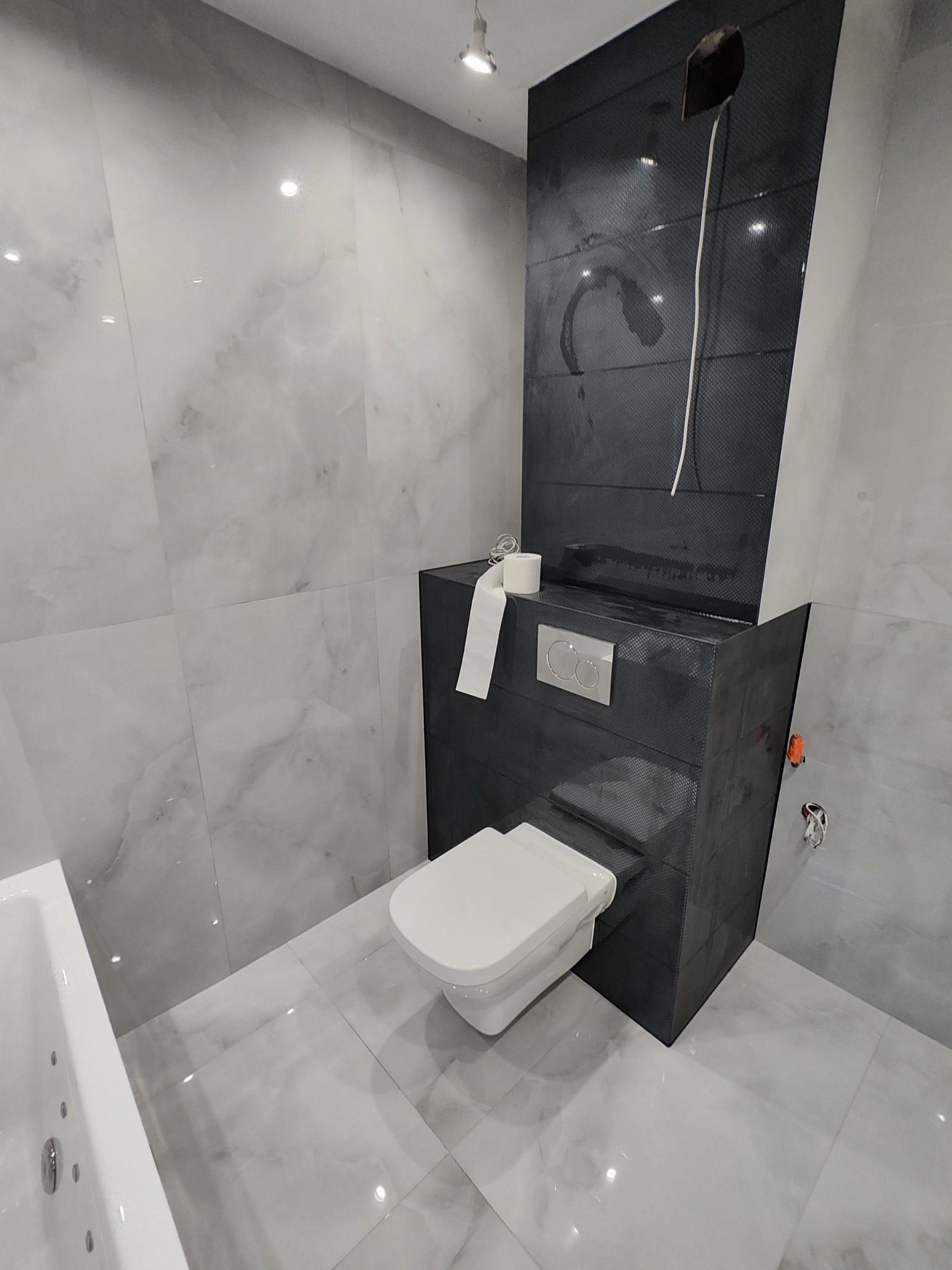 Wykończona łazienka z podwieszanym WC w obudowie z ciemnych płytek i marmurowymi płytkami na ścianach. Widoczna wanna z hydromasażem.