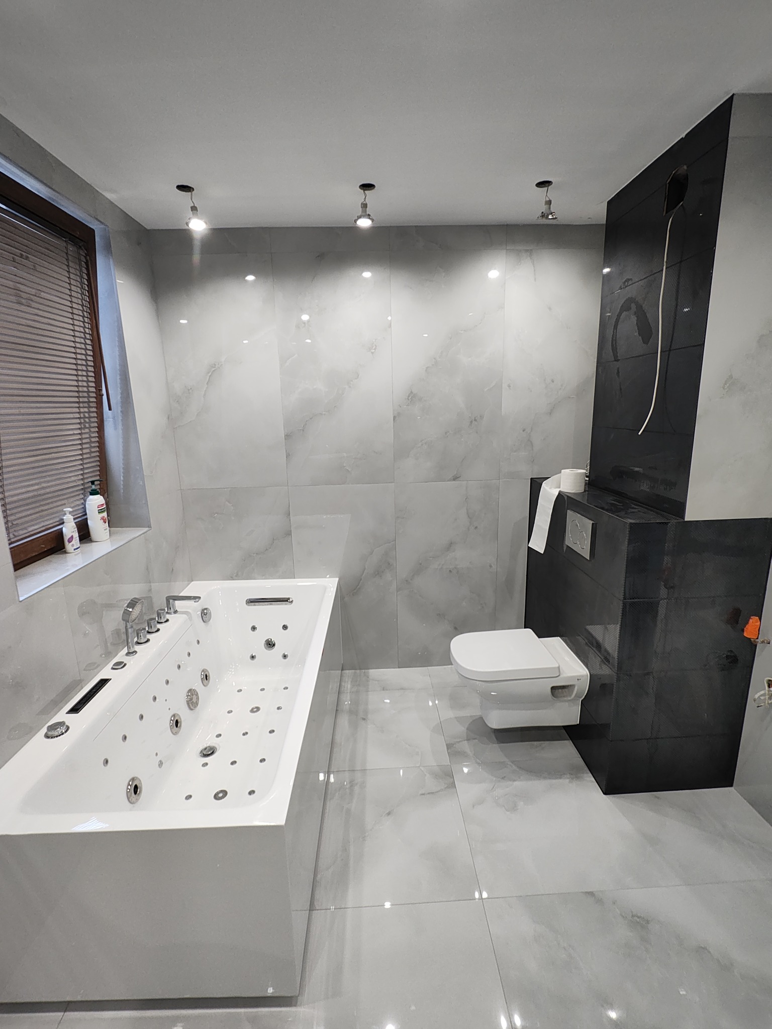 Wykończona, nowoczesna łazienka z białą wanną jacuzzi, podwieszaną toaletą i szarymi płytkami imitującymi marmur. Widoczne punktowe oświetlenie i okno z żaluzjami.