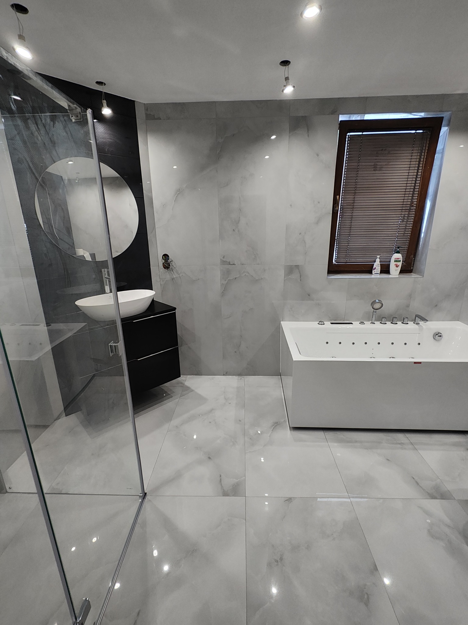 Elegancka łazienka z wanną jacuzzi, umywalką nablatową na czarnej szafce i kabiną prysznicową. Marmurowe płytki na ścianach i podłodze, okno z żaluzjami.