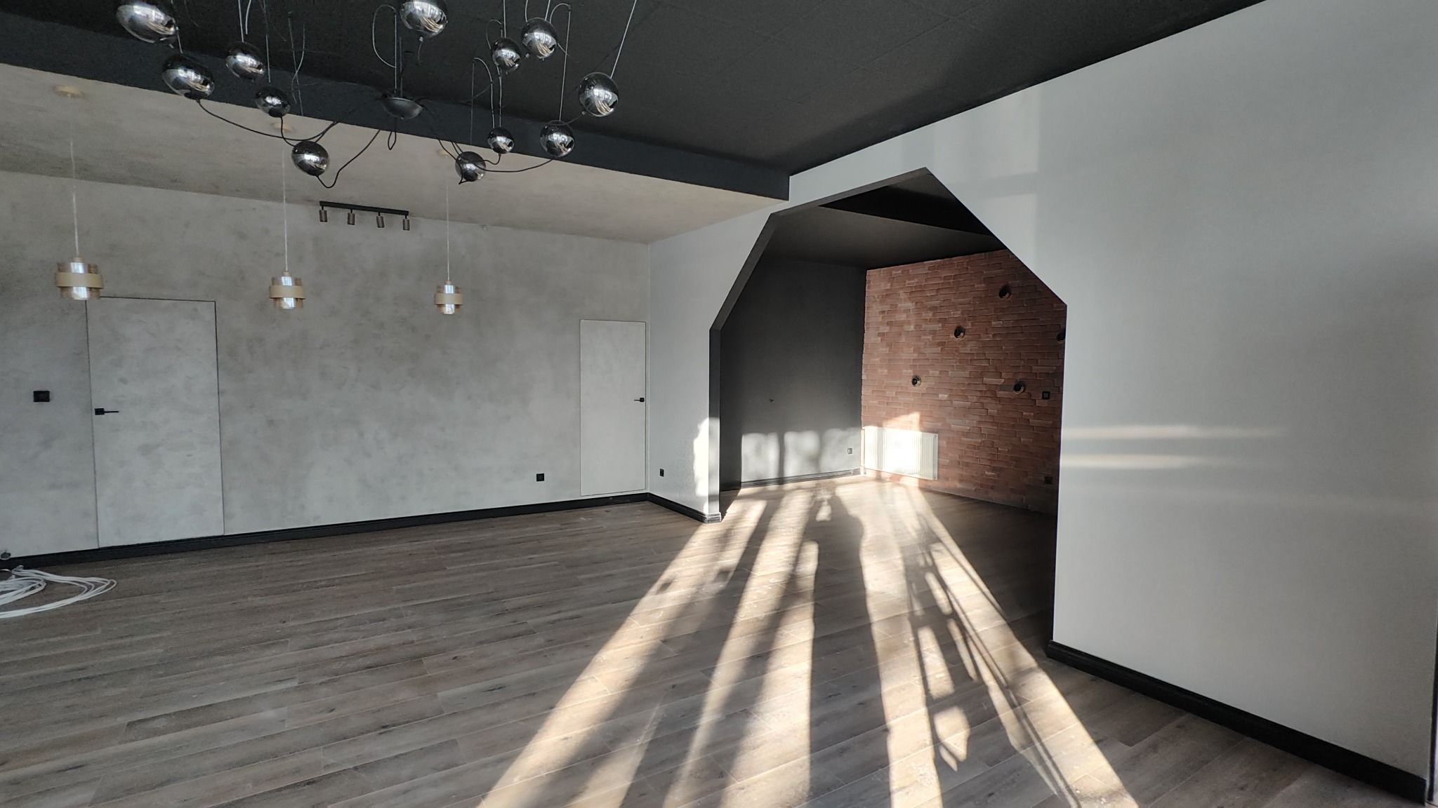 Nowoczesne wnętrze po remoncie: drewnopodobna podłoga, ściany w odcieniach szarości i bieli, ceglana ściana z grzejnikiem, designerskie lampy sufitowe.