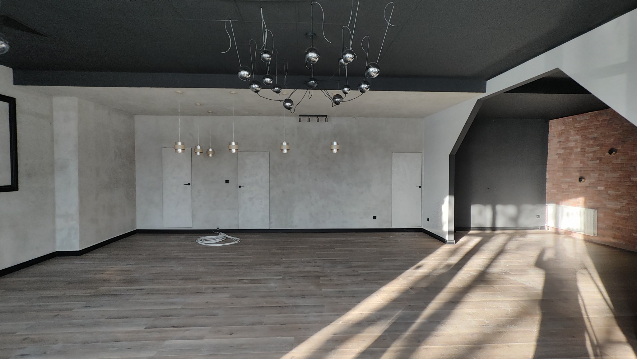 Wykończone wnętrze w stylu loft: drewniana podłoga, szare ściany z efektem betonu, czarny sufit i designerska lampa. Widoczne trzy ukryte drzwi i fragment ściany z cegły.