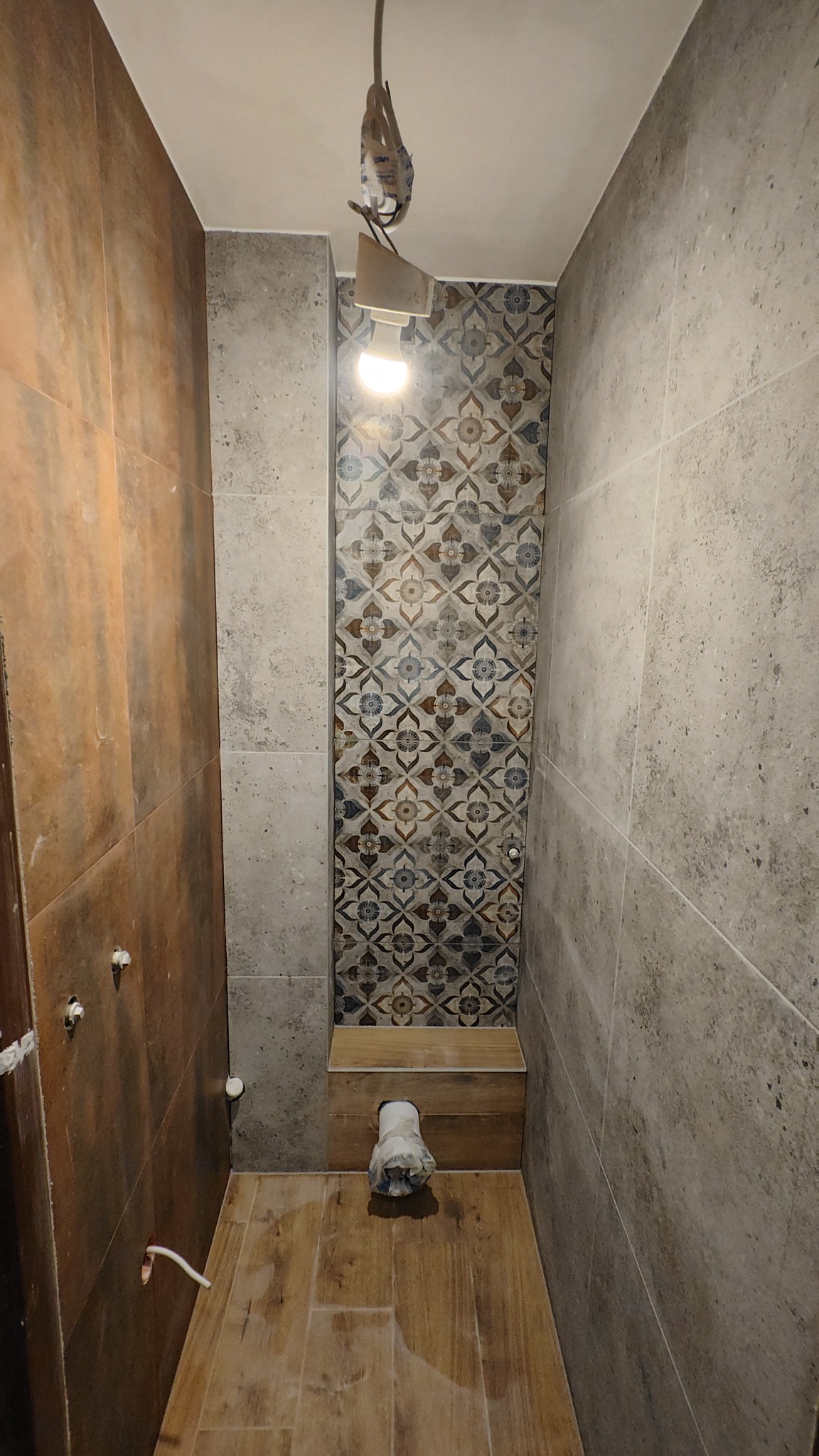 Wnętrze łazienki w trakcie wykańczania: brązowe i szare płytki na ścianach, drewnopodobna podłoga, dekoracyjna wnęka z wzorzystymi płytkami, widoczne instalacje.