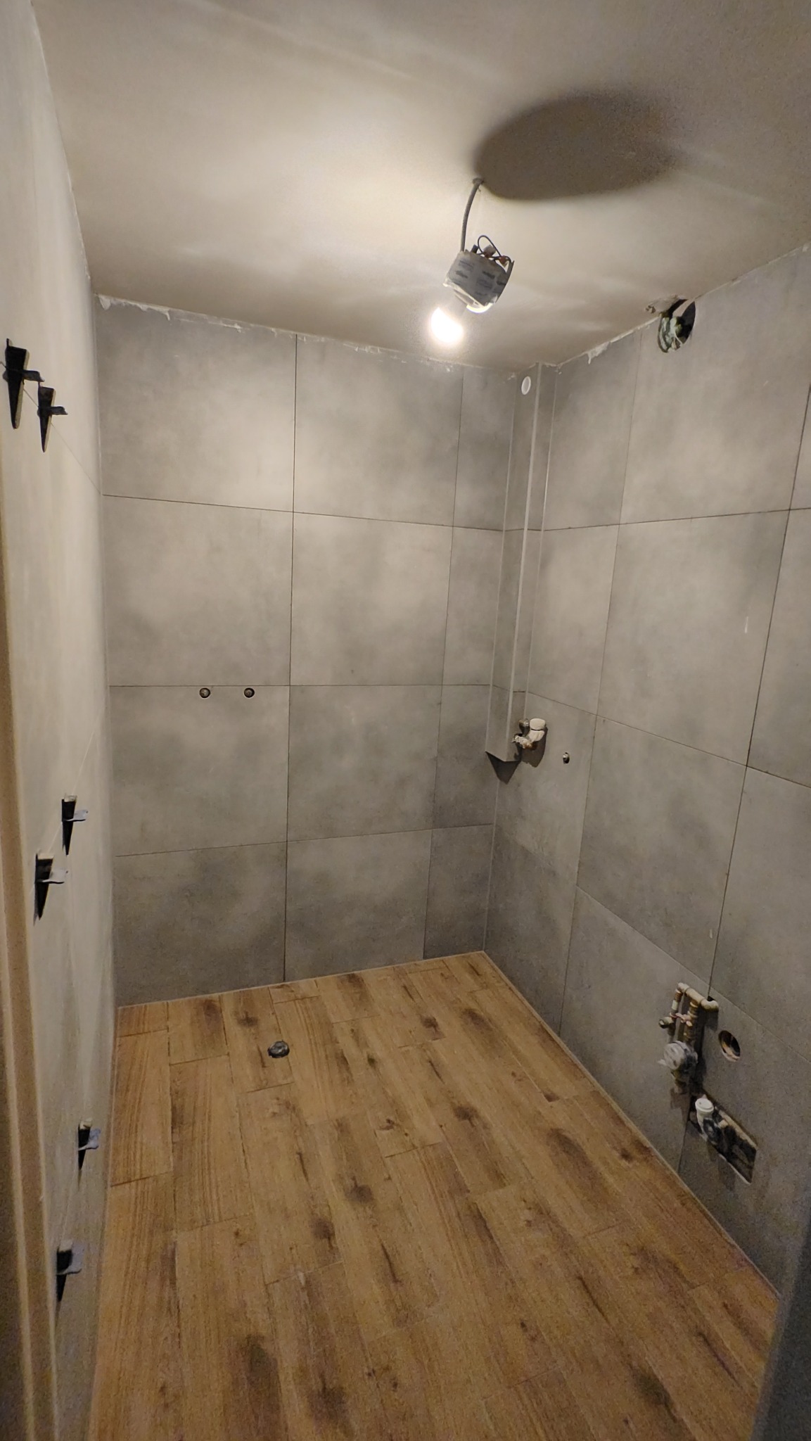 Szara łazienka w trakcie wykańczania: ułożone płytki podłogowe imitujące drewno, szare płytki ścienne z otworami na armaturę, widoczne krzyżyki glazurnicze.