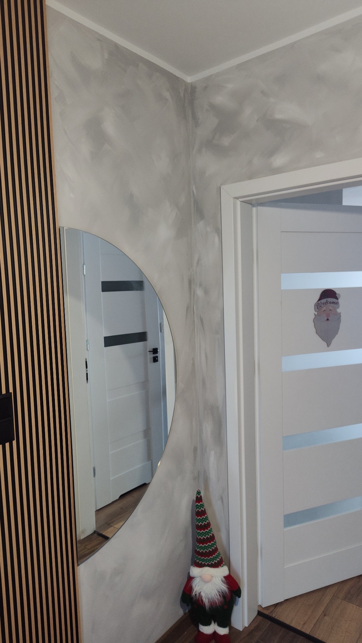 Ściana z efektem dekoracyjnym w odcieniach szarości, drewniane lamele, okrągłe lustro i świąteczny skrzat. Widoczny fragment białych drzwi z dekoracją.