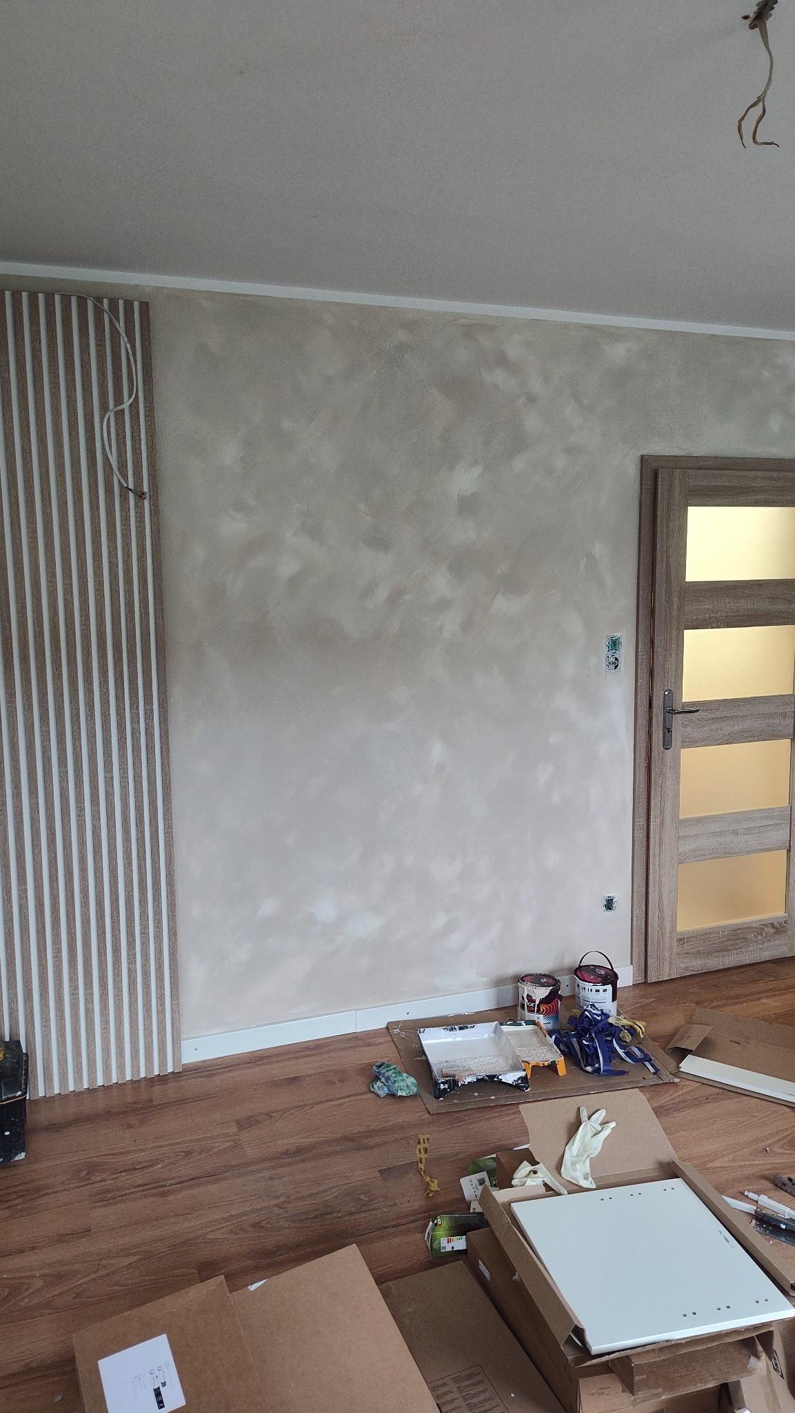 Ściana z efektem dekoracyjnym w beżach, listwy ścienne, drzwi z przeszkleniami. Na podłodze akcesoria malarskie i kartony. Widoczne gniazdka elektryczne.