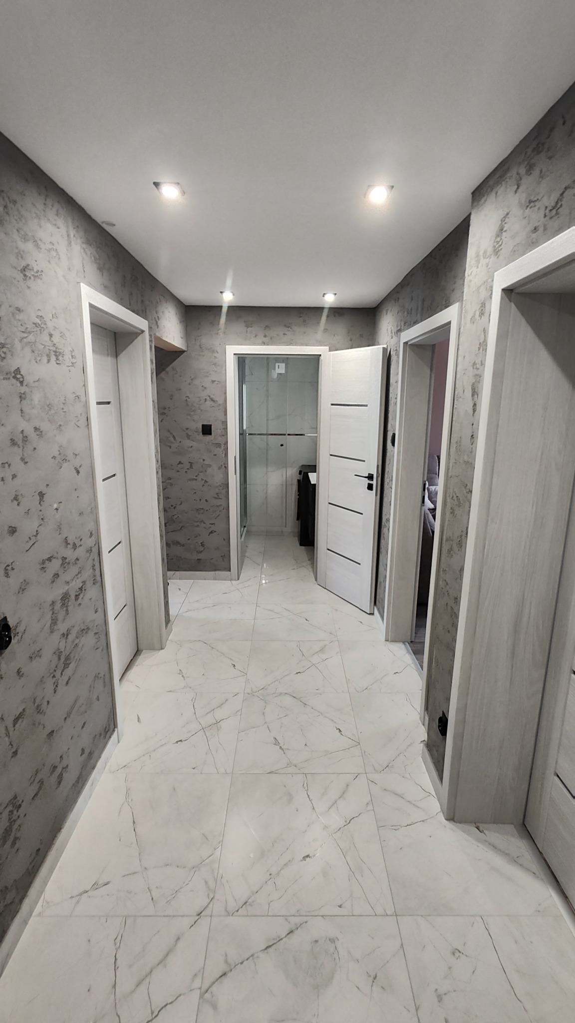 Wykończony korytarz z marmurową podłogą i dekoracyjną szarą ścianą. Widoczne trzy białe drzwi z czarnymi klamkami i oświetlenie sufitowe.