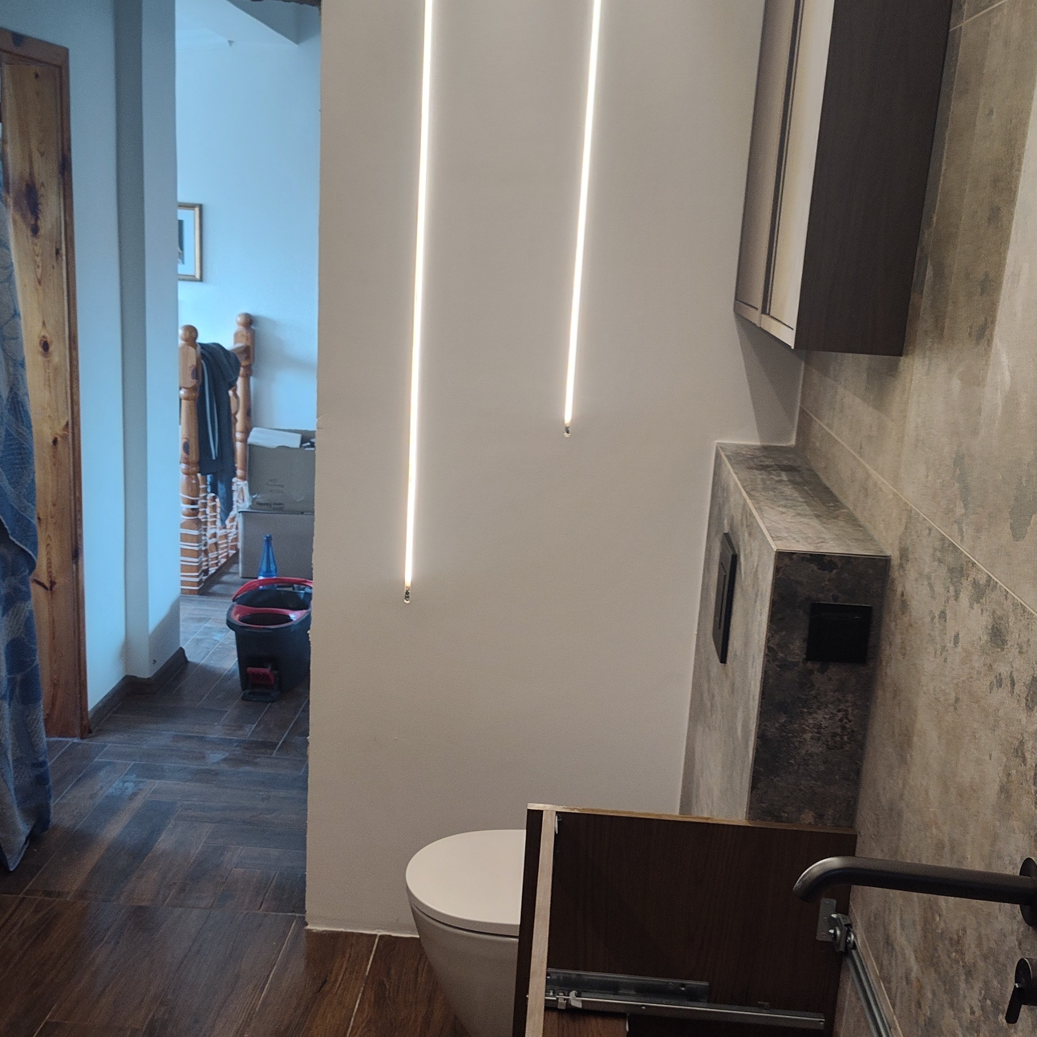 Nowoczesna łazienka po remoncie: podświetlane pionowe linie na ścianie, umywalka z szafką, płytki imitujące kamień, drewnopodobna podłoga.