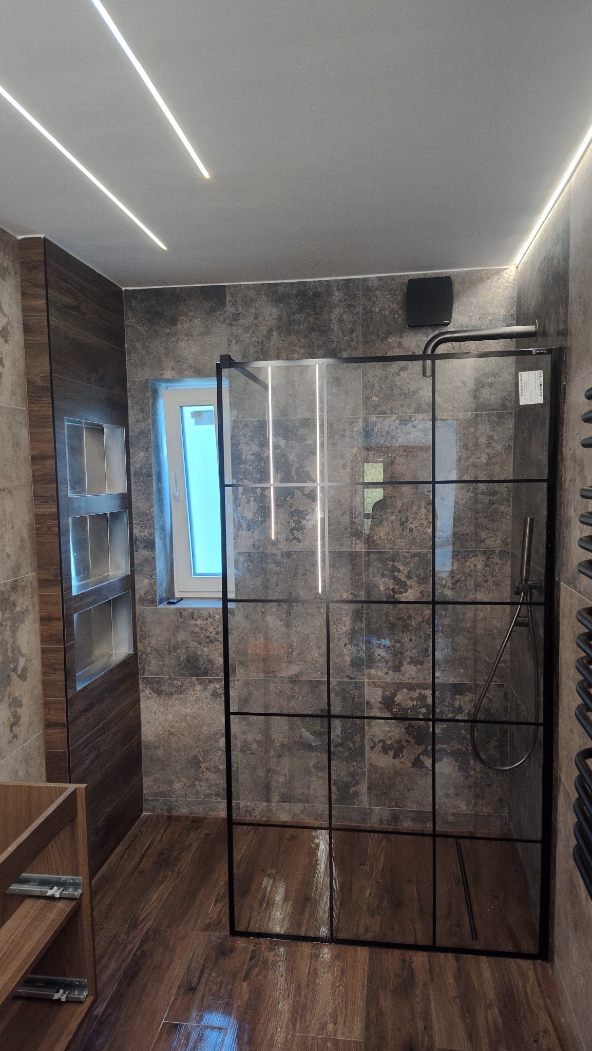 Nowoczesna łazienka z kabiną prysznicową typu walk-in z czarnymi szprosami, drewnopodobnymi płytkami i oświetleniem LED. Widok na okno i wnęki w ścianie.