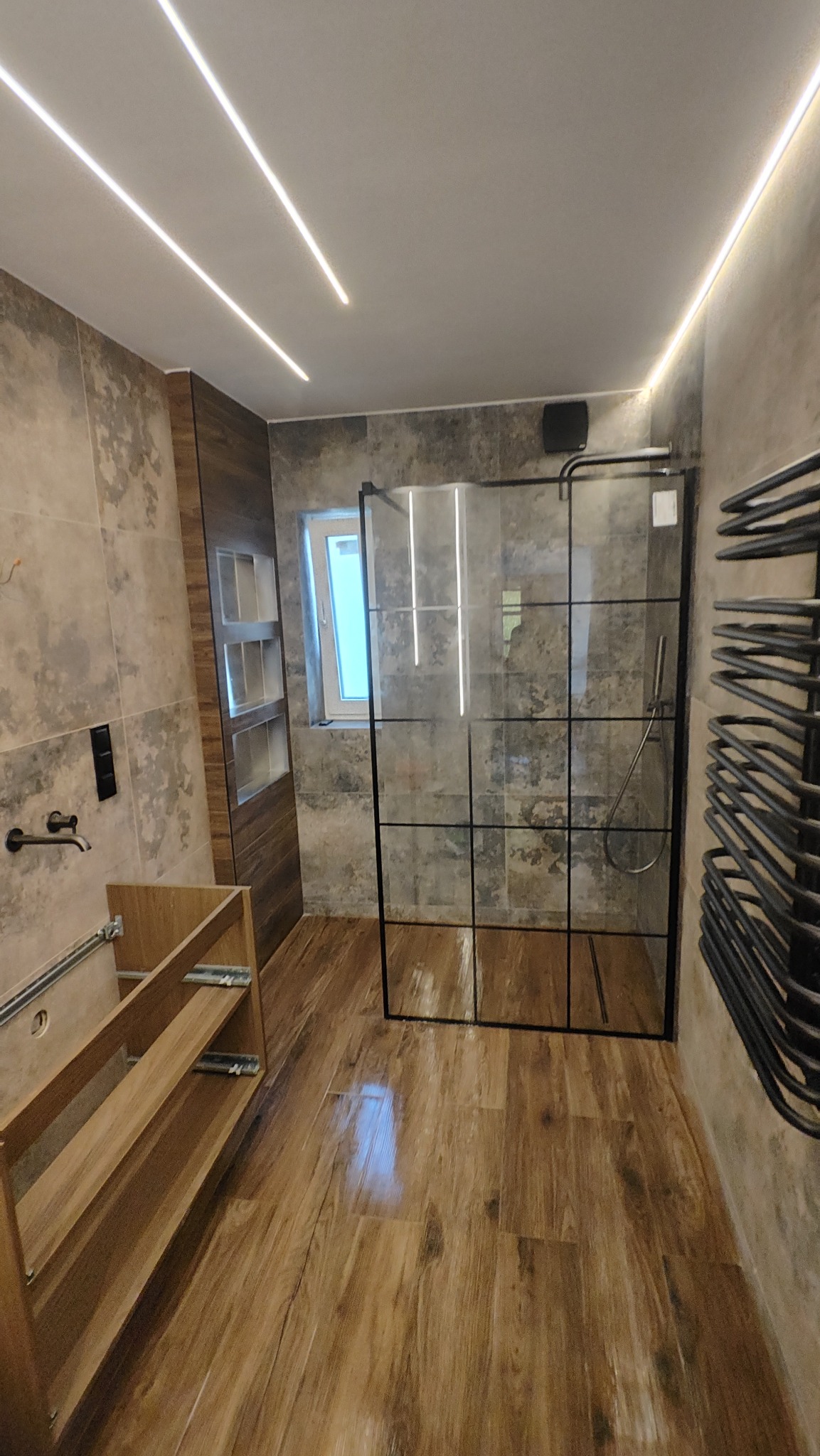 Nowoczesna łazienka z kabiną typu walk-in, drewnopodobnymi płytkami na podłodze i ścianach, oraz podświetleniem LED. Widoczny fragment szafki pod umywalkę.