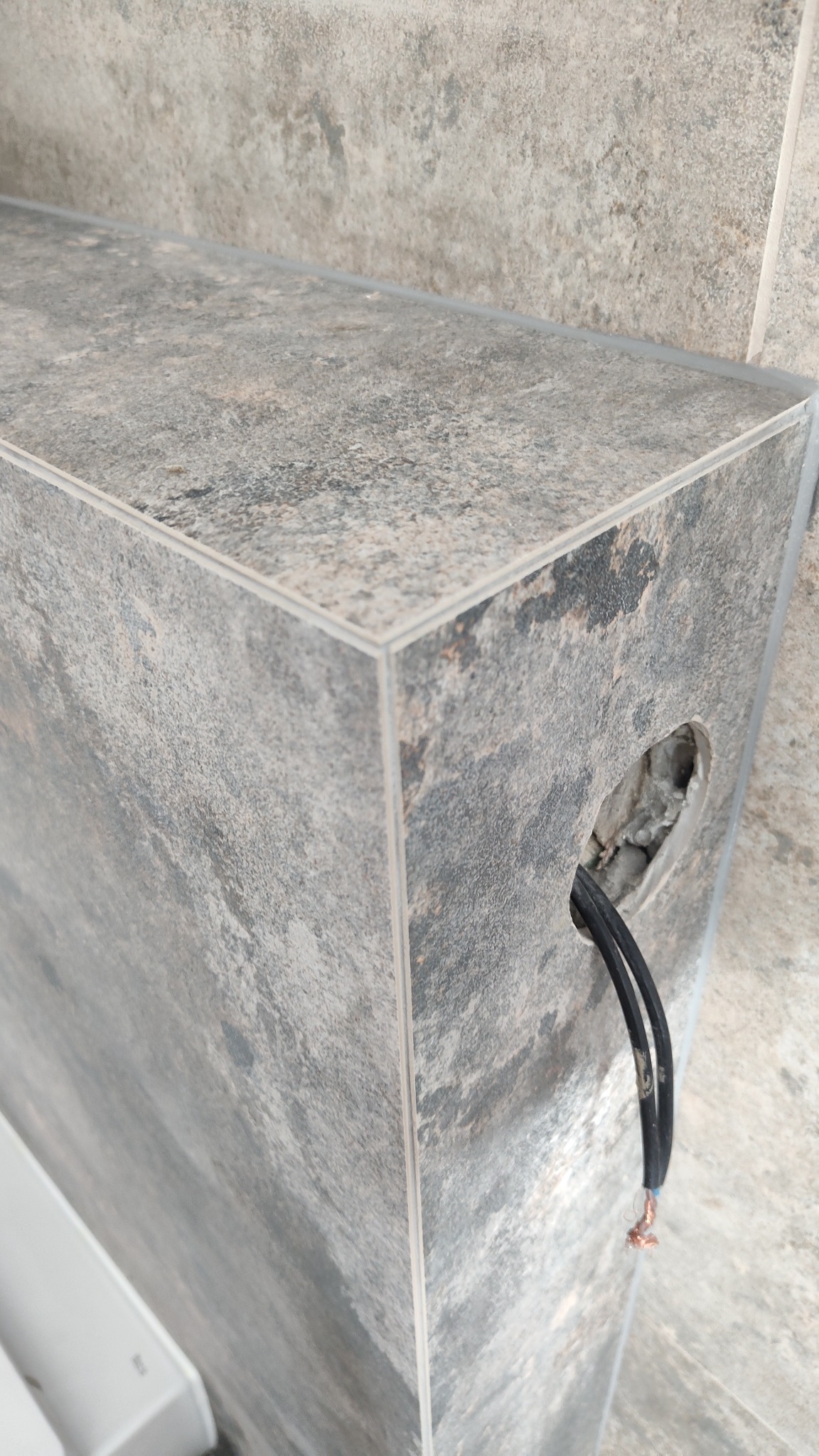 Szare płytki imitujące kamień, ułożone pod kątem, z otworem na przewody elektryczne. Widoczne kable wystające ze ściany. Precyzyjne wykończenie narożników.