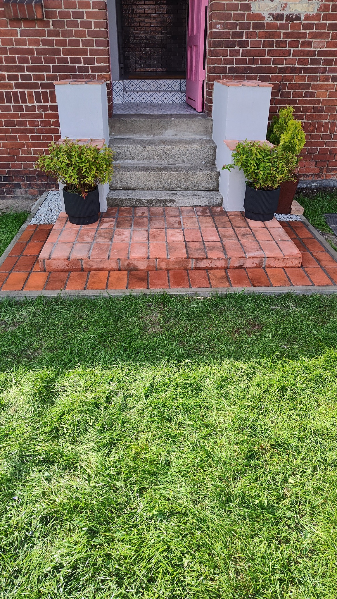 Wejście do domu z ceglanym podestem i schodami, ozdobione roślinami w donicach. W tle różowe drzwi i wzorzyste płytki na podeście.