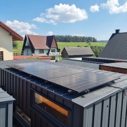 ELMAX SPÓŁKA Z OGRANICZONĄ ODPOWIEDZIALNOŚCIĄ - Panele słoneczne zamontowane na dachu nowoczesnego, modułowego domu z szarej blachy trapezowej, widok z góry na tle zielonego krajobrazu i błękitnego nieba z chmurami.