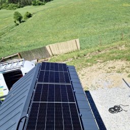 ELMAX SPÓŁKA Z OGRANICZONĄ ODPOWIEDZIALNOŚCIĄ - Dwa panele słoneczne zamontowane na szarym dachu blaszanym, widok z góry na zielone wzgórze i częściowo widoczny samochód dostawczy z drabiną.