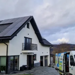 Biały, dwupiętrowy dom z czarnymi panelami na dachu, zaparkowany obok biały van z logo firmy na tle pochmurnego nieba i pag&oacute;rkowatego krajobrazu.