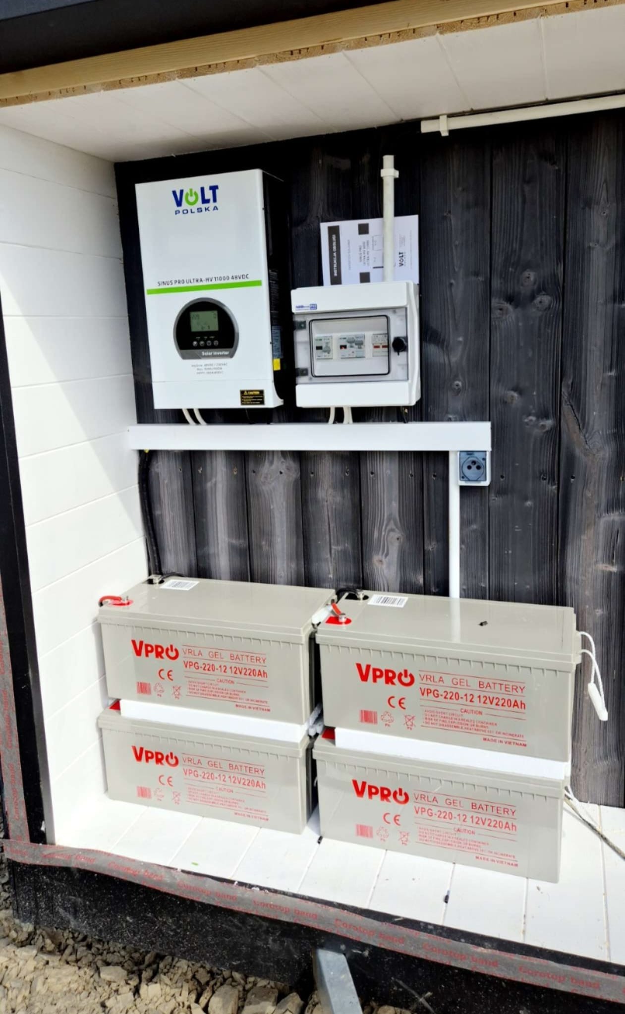 Szafka z zainstalowanym inwerterem Volt Polska, rozdzielnicą elektryczną i czterema akumulatorami VPRO VRLA GEL 12V220Ah, zamontowana na drewnianej ścianie.