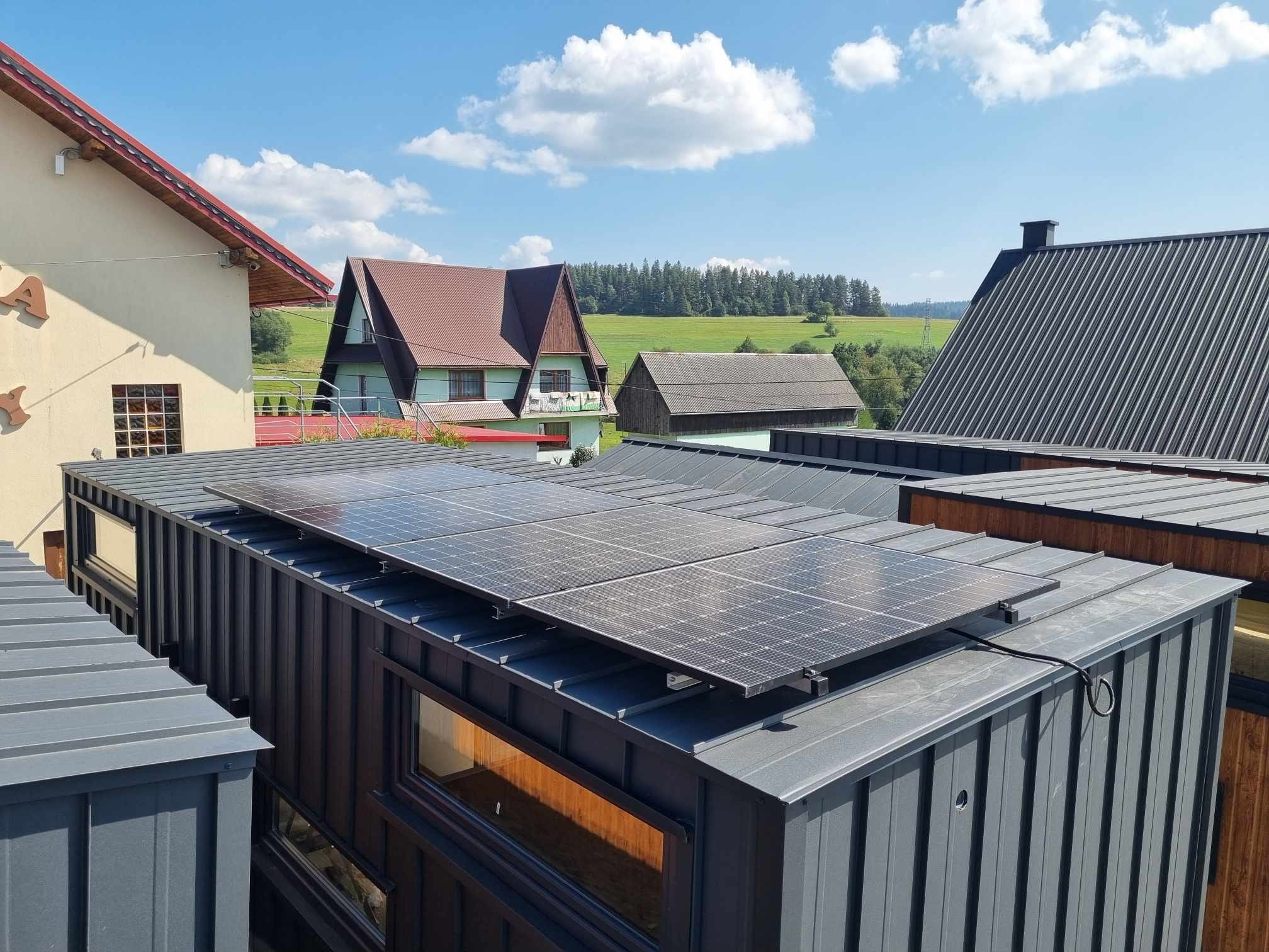 Panele słoneczne zamontowane na dachu nowoczesnego, modułowego domu z szarej blachy trapezowej, widok z góry na tle zielonego krajobrazu i błękitnego nieba z chmurami.