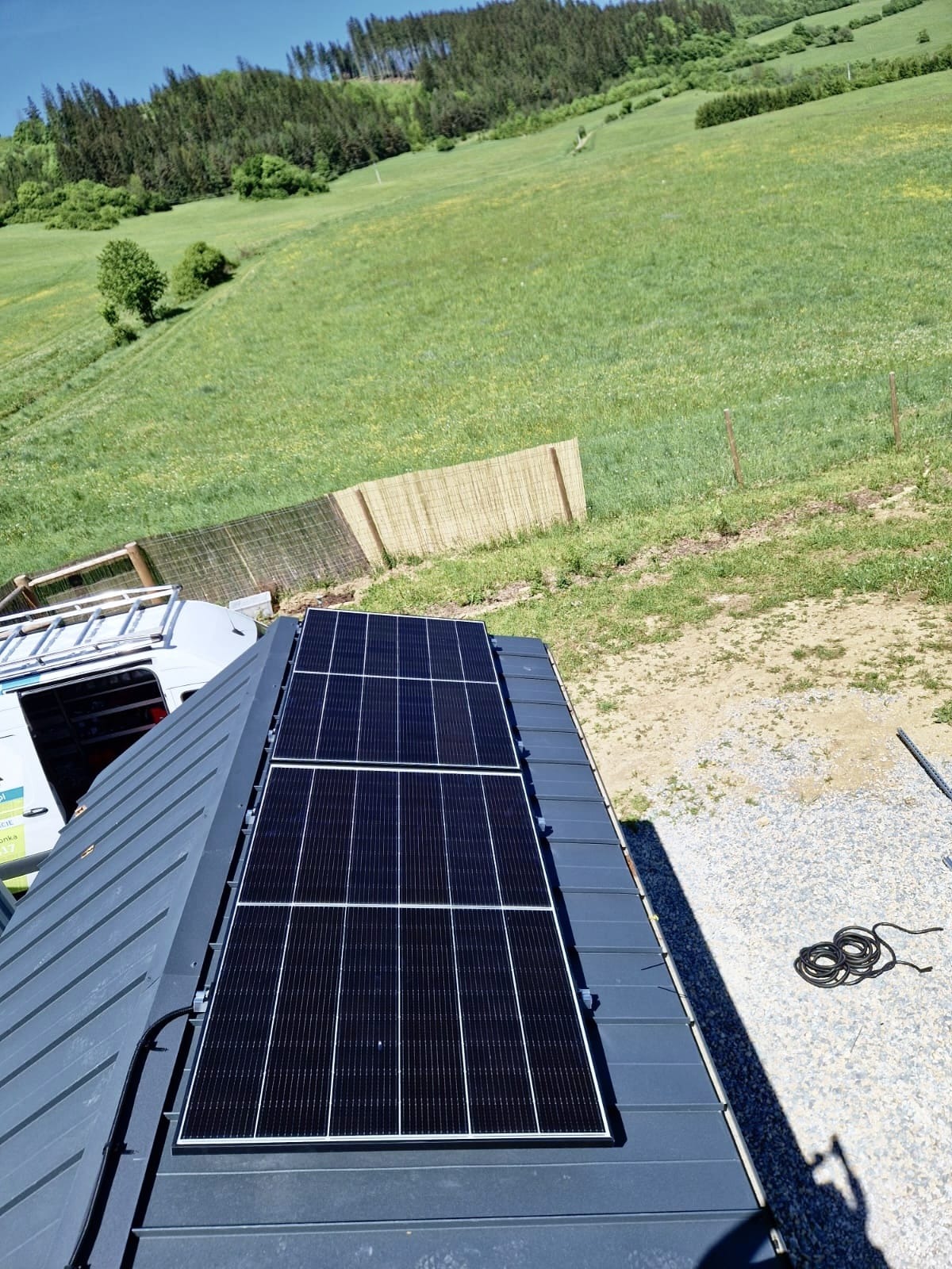 Dwa panele słoneczne zamontowane na szarym dachu blaszanym, widok z góry na zielone wzgórze i częściowo widoczny samochód dostawczy z drabiną.