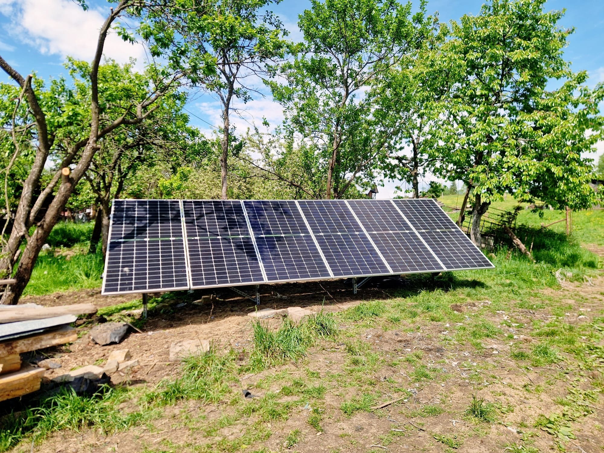 Rząd paneli słonecznych ustawionych na metalowej konstrukcji na trawiastym terenie, otoczony drzewami owocowymi w słoneczny dzień.