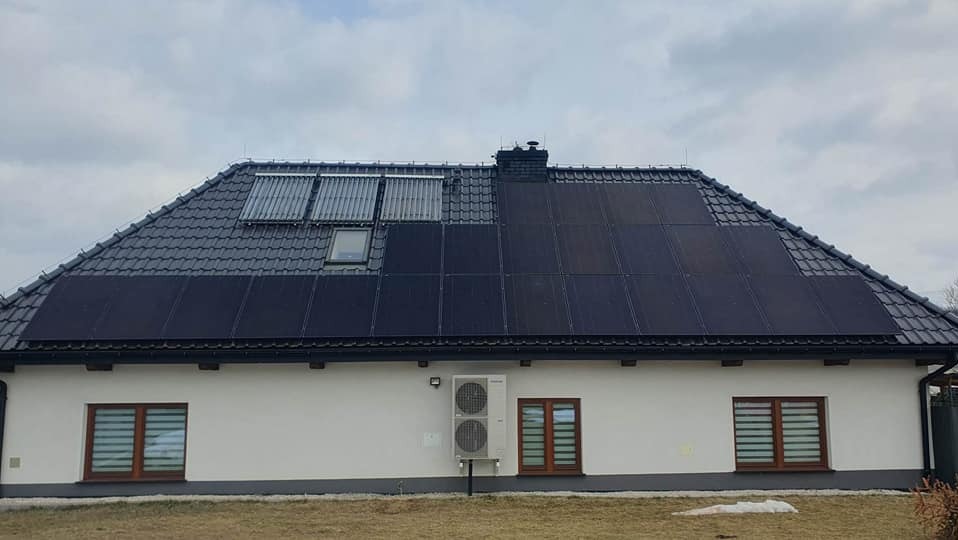 Dom jednorodzinny z zainstalowanymi panelami słonecznymi na dachu, połączonymi z kolektorami słonecznymi; widoczna jednostka zewnętrzna klimatyzacji na ścianie budynku.