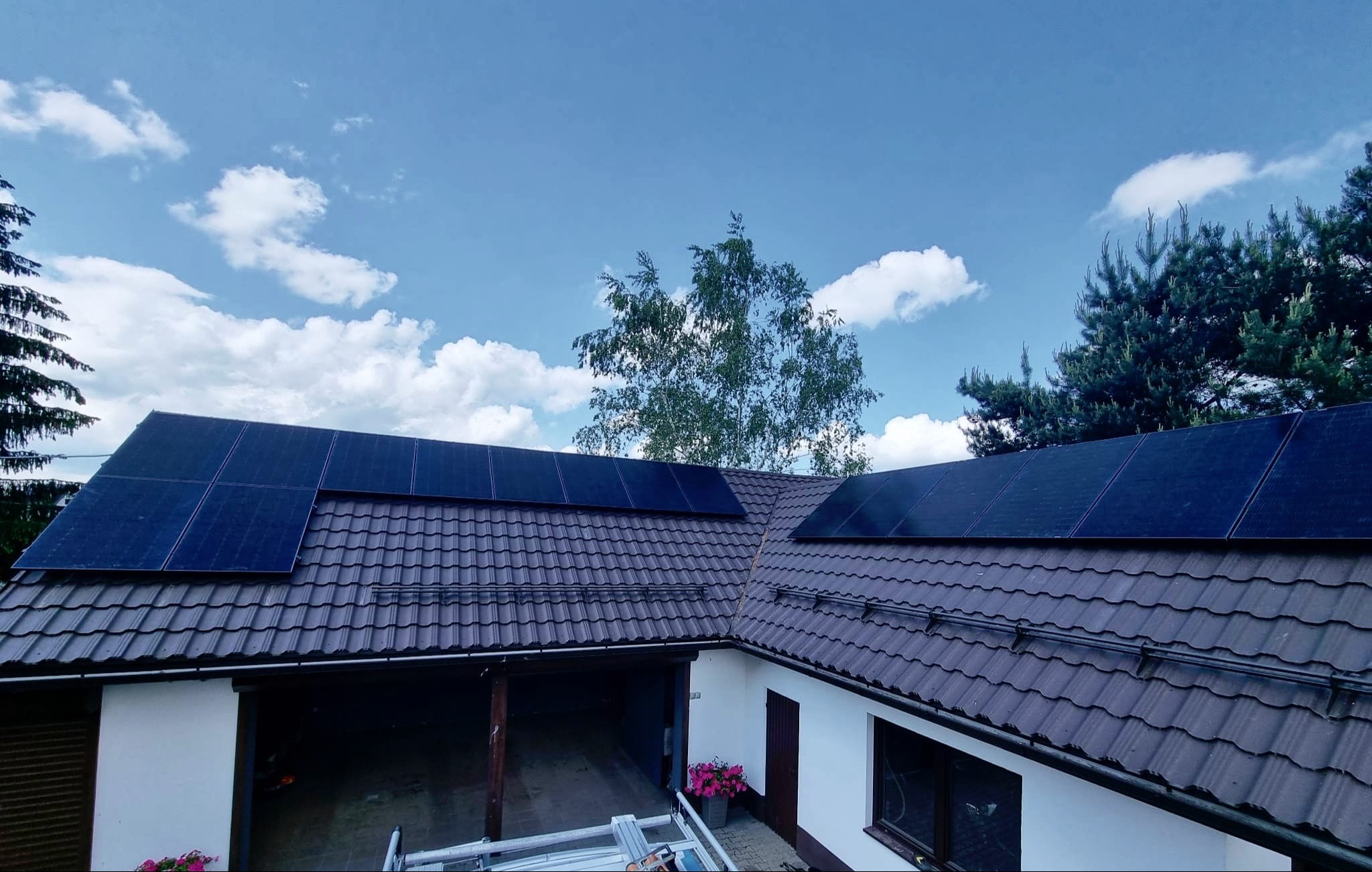 Panele słoneczne na brązowym dachu domu jednorodzinnego, widok z góry na tle błękitnego nieba z białymi chmurami i zieleni drzew.