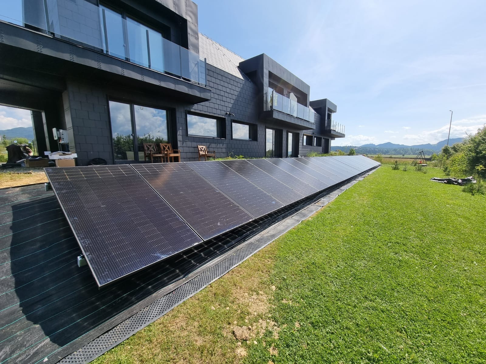 Rząd paneli fotowoltaicznych, zainstalowanych przy nowoczesnym, dwupiętrowym domu z elewacją z łupka, na trawniku z widokiem na górzysty krajobraz.