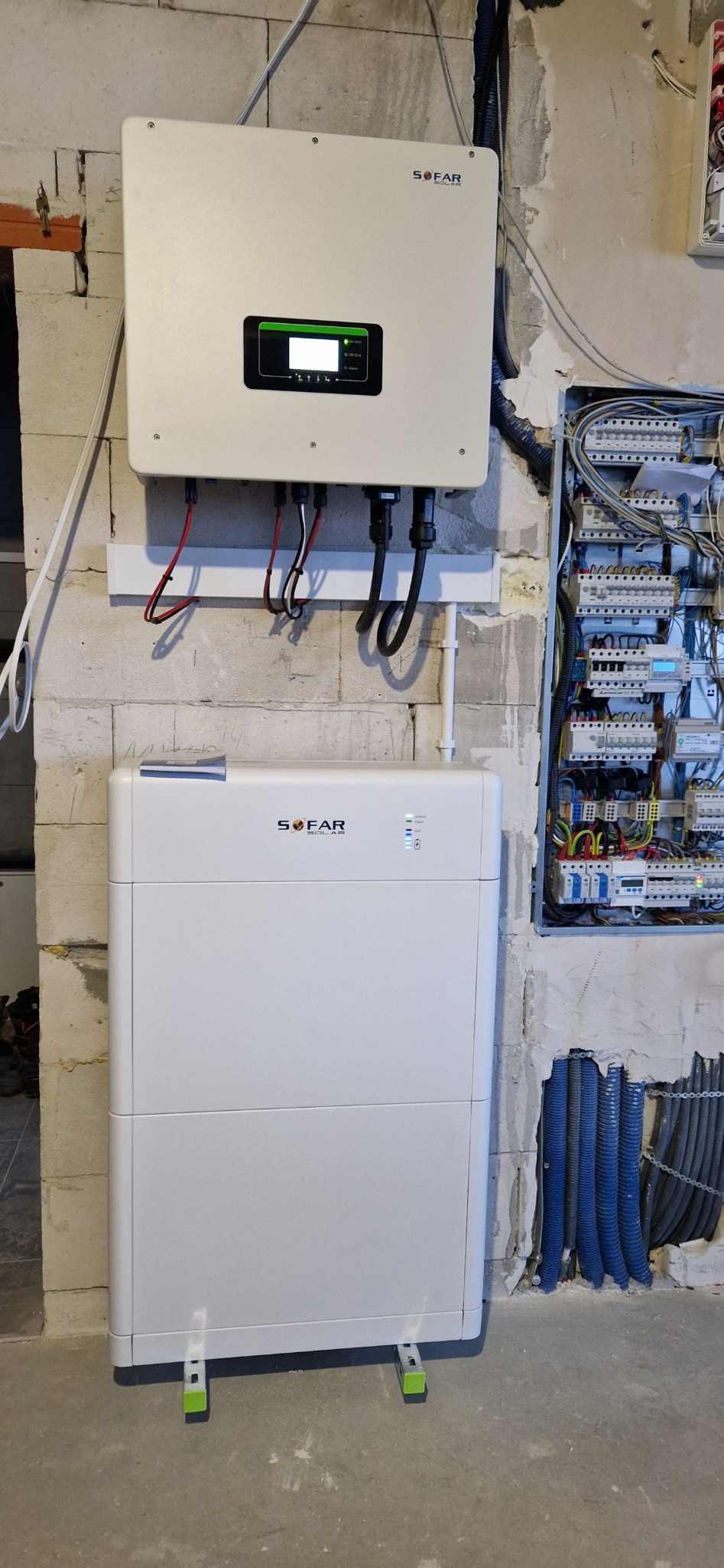 Biały inwerter i magazyn energii marki Sofar zamontowane na surowej ścianie z betonu komórkowego, obok skrzynka elektryczna z licznymi przewodami.