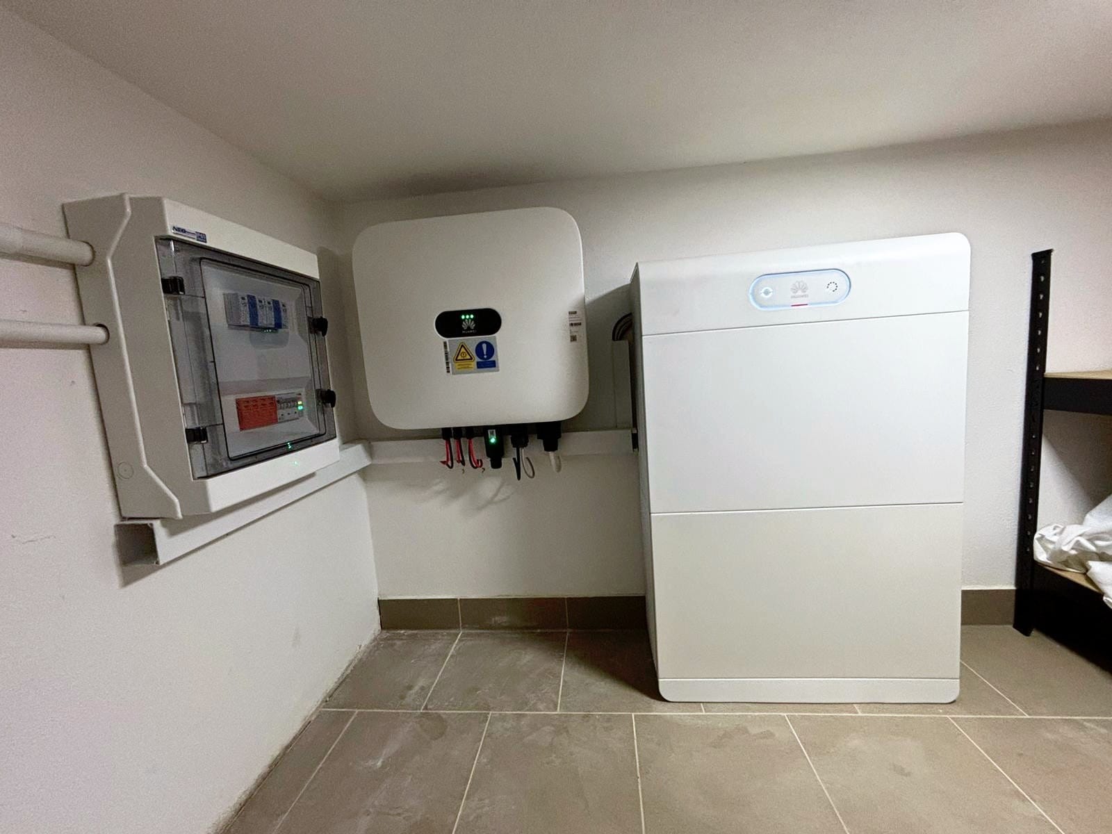 Biała skrzynka rozdzielcza, inwerter i akumulator energii Huawei zamontowane na ścianie w pomieszczeniu z szarą podłogą z płytek.