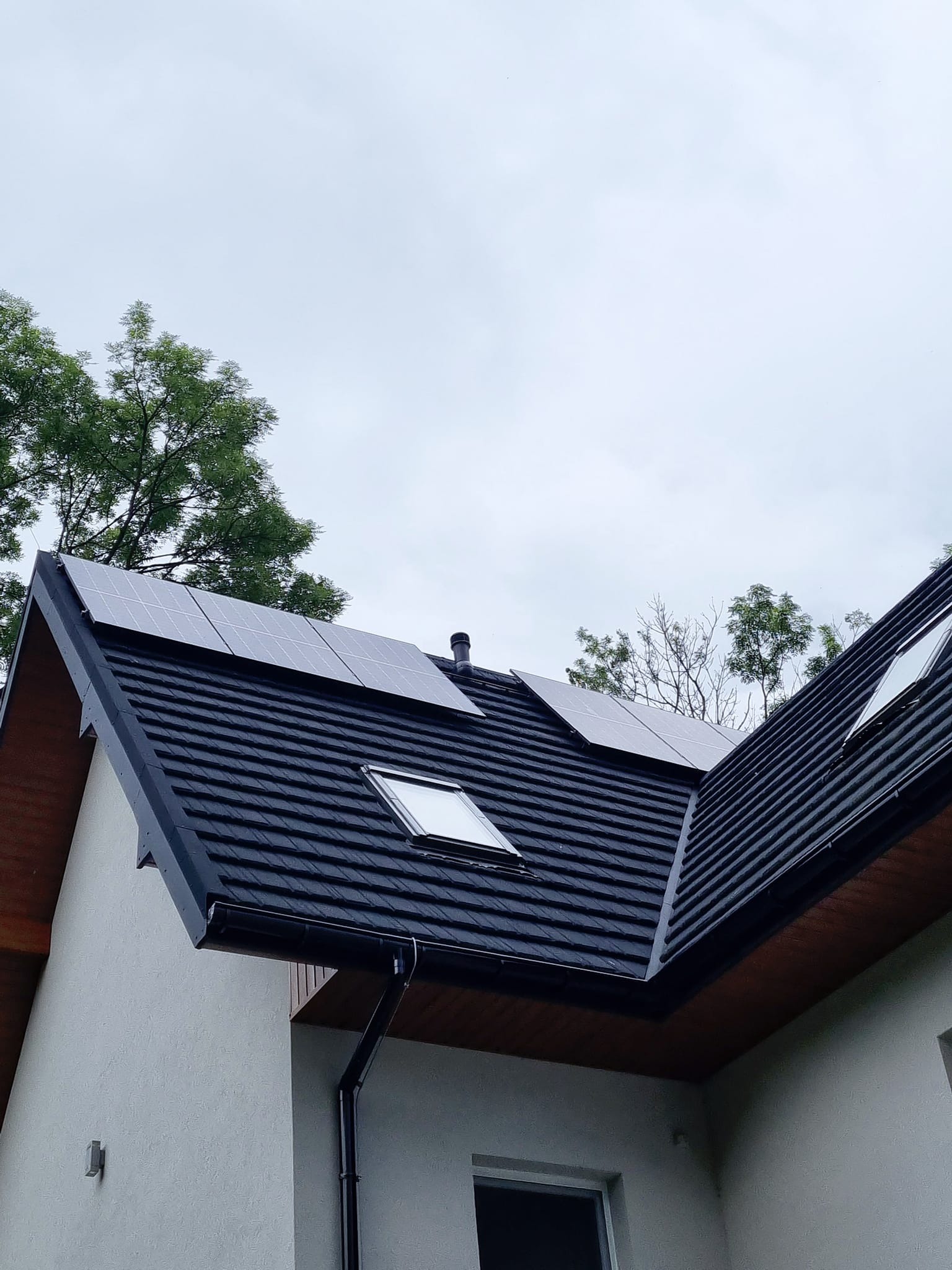 Panele słoneczne zamontowane na dachu domu z czarną dachówką, widoczne okno dachowe i fragment systemu rynnowego.