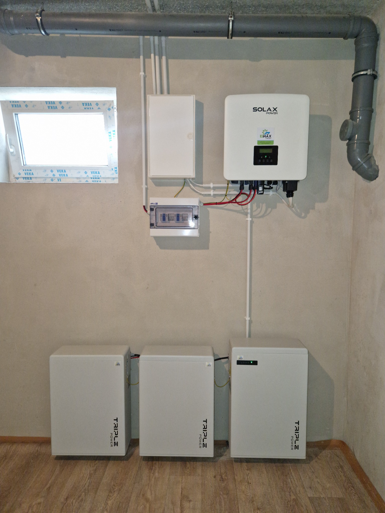 Instalacja domowa z trzema białymi magazynami energii Triple Power, inwerterem Solax Power, rozdzielnicą elektryczną i okablowaniem, widoczne na tle surowej ściany z oknem.