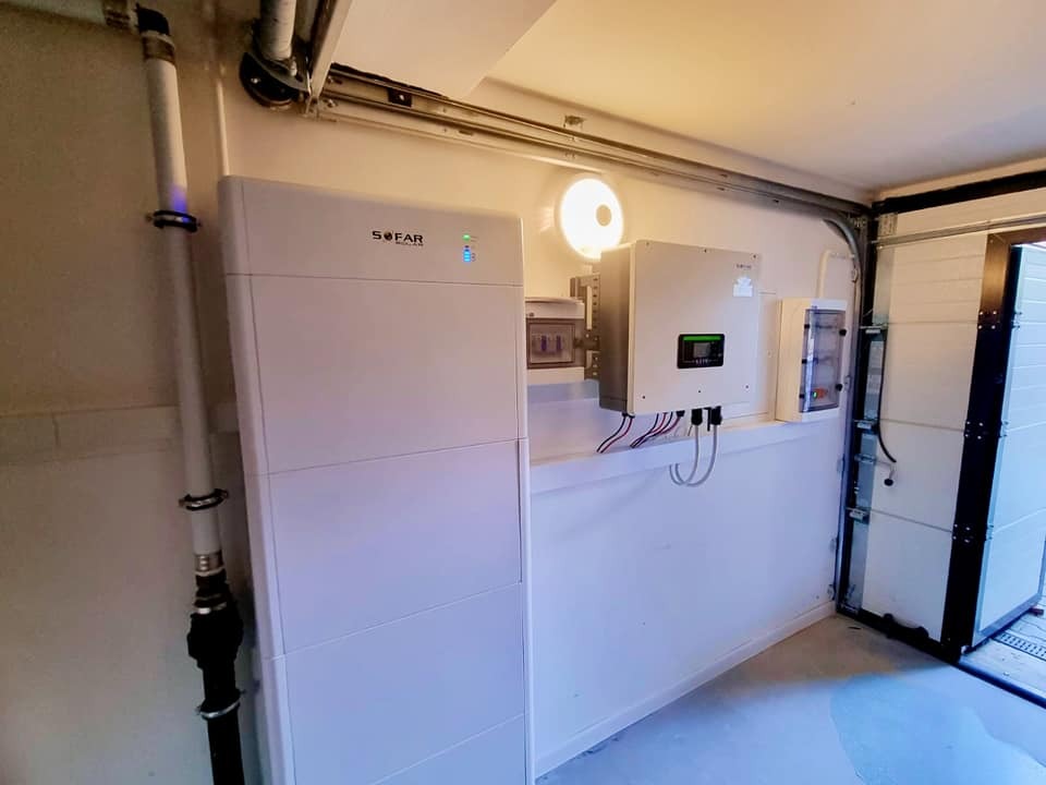 Biały magazyn energii marki Sofar, zamontowany w garażu obok falownika i skrzynki elektrycznej, z widocznymi przewodami i fragmentem otwartej bramy garażowej.