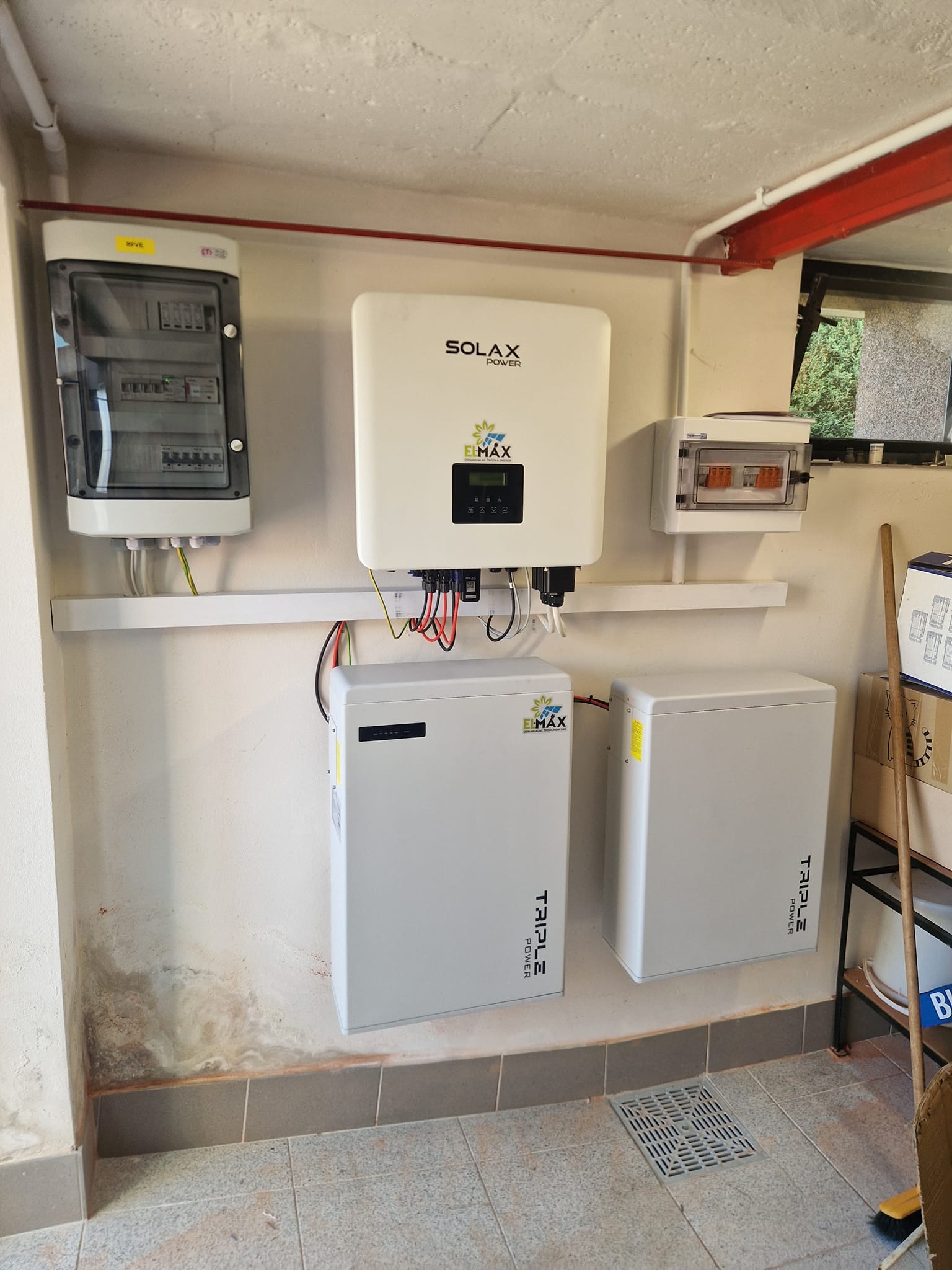 Białe akumulatory energii Elmax Triple Power i falownik Solax Power zamontowane na ścianie w pomieszczeniu gospodarczym, widoczna skrzynka elektryczna i fragment okna.