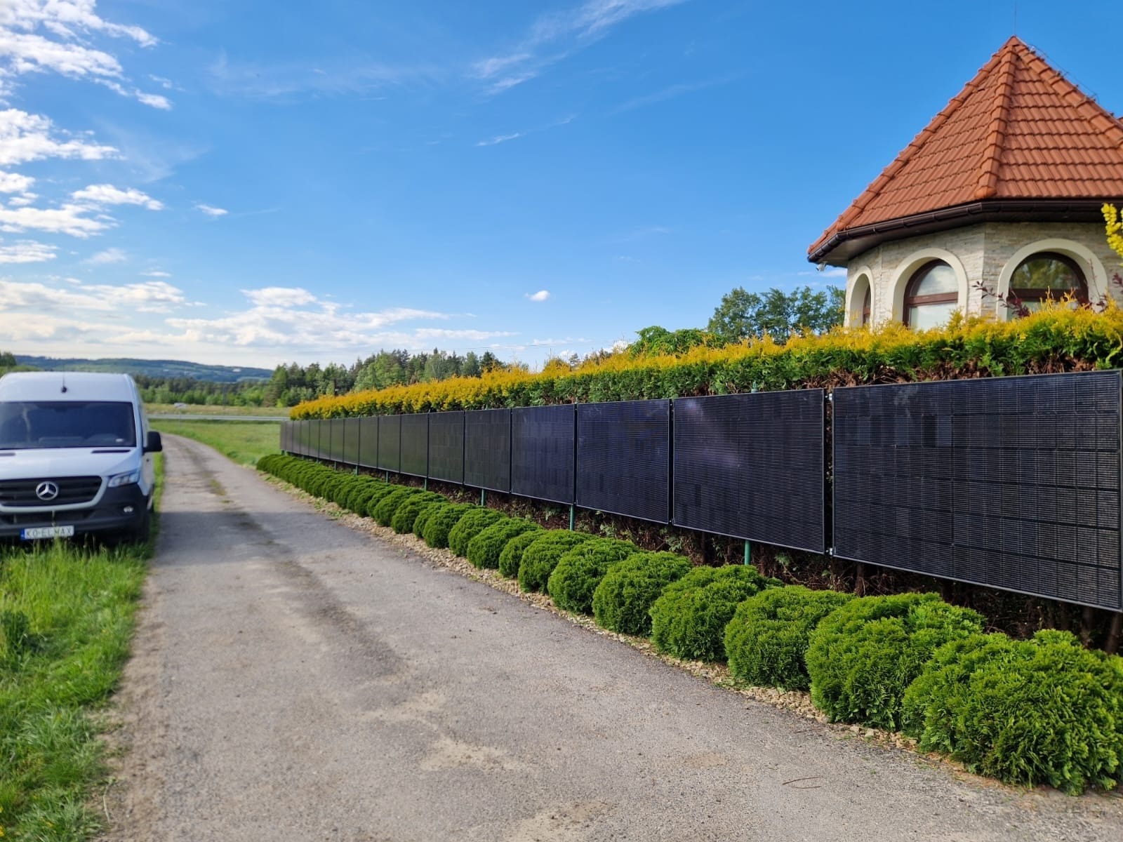Długa instalacja paneli na ogrodzeniu, widoczny biały samochód dostawczy Mercedes i fragment budynku z czerwoną dachówką w tle, błękitne niebo.