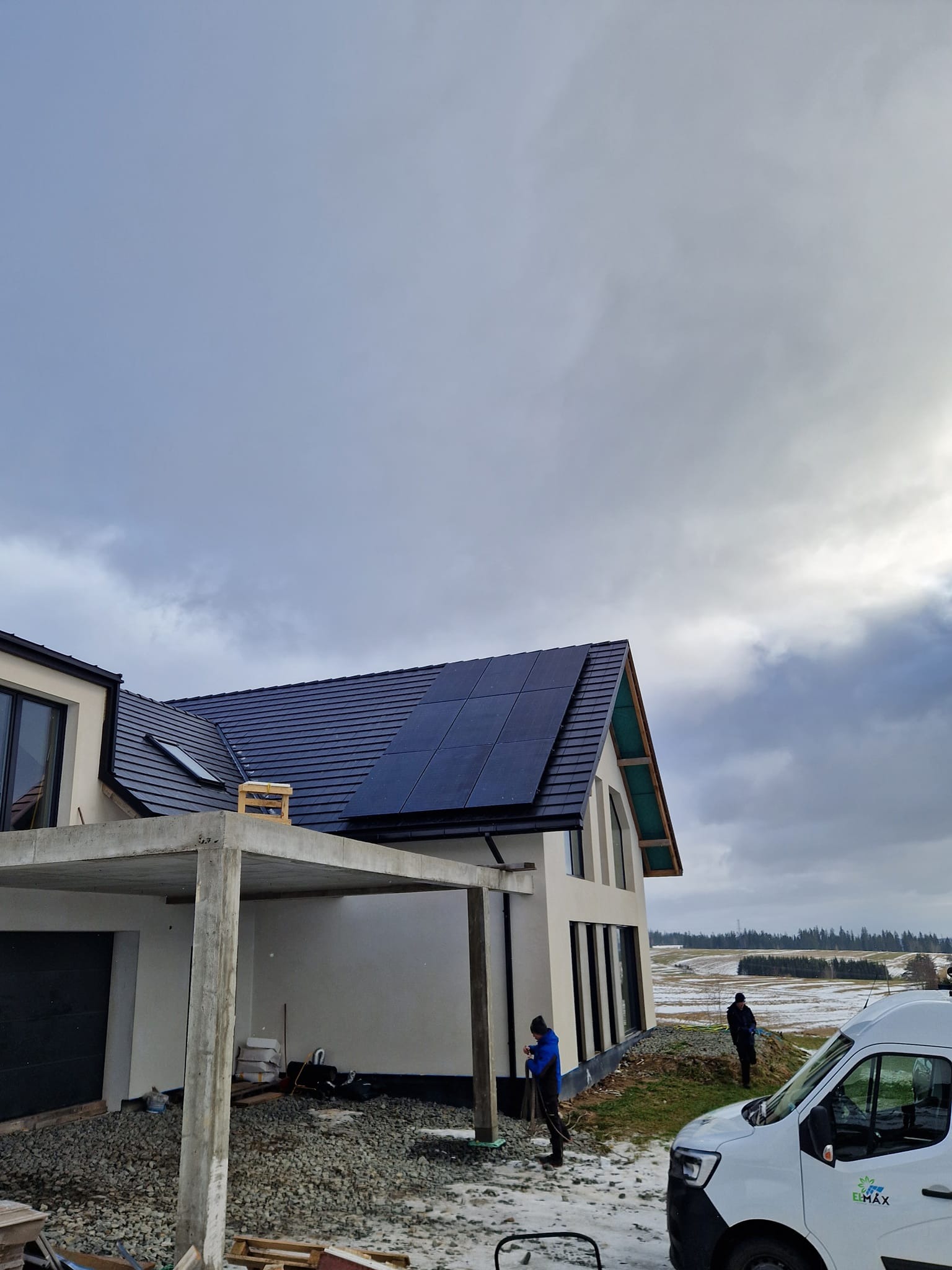 Montaż paneli słonecznych na dachu nowoczesnego domu z widocznymi pracownikami i samochodem firmy na tle zimowego krajobrazu.