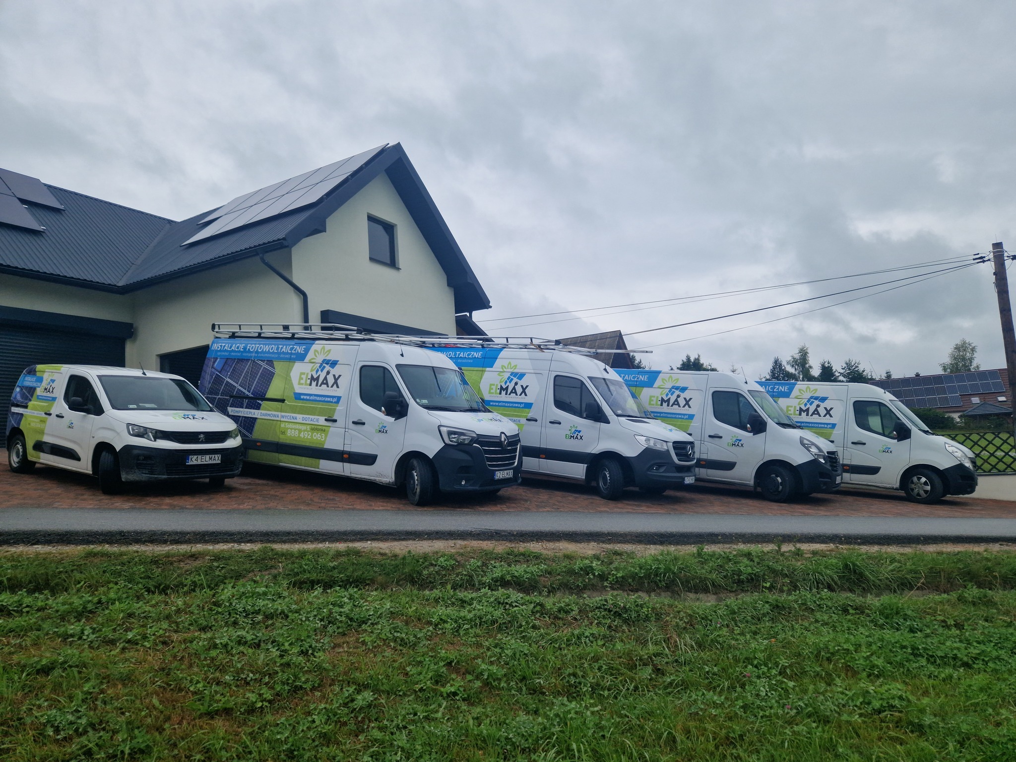 Flota pięciu białych samochodów dostawczych z logo firmy i reklamą instalacji paneli słonecznych zaparkowana przed domem z zamontowaną instalacją fotowoltaiczną.