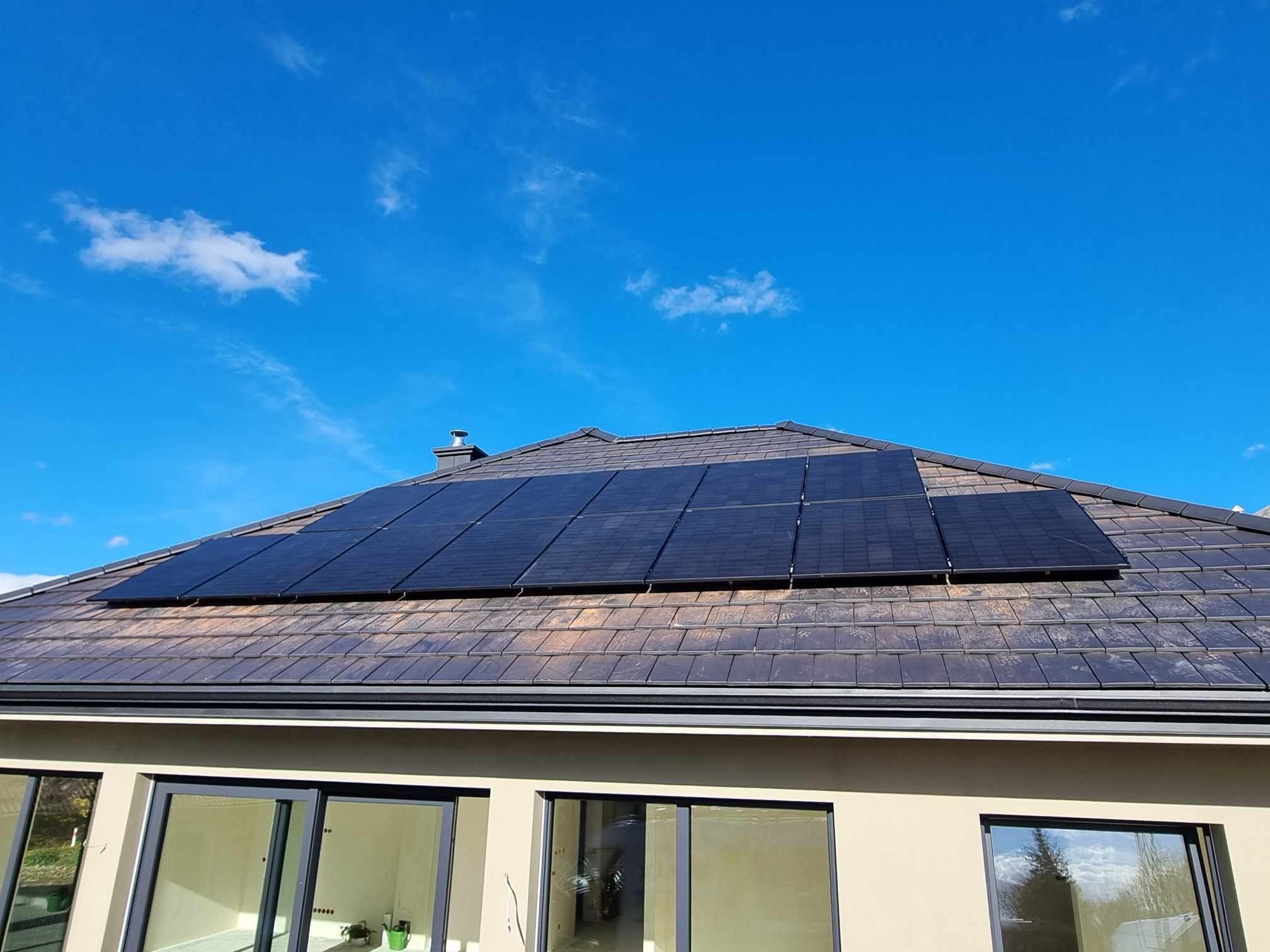 Panele fotowoltaiczne na dachu domu z dachówką bitumiczną, widoczne okna z przesuwanymi drzwiami i błękitne niebo z chmurami w tle.