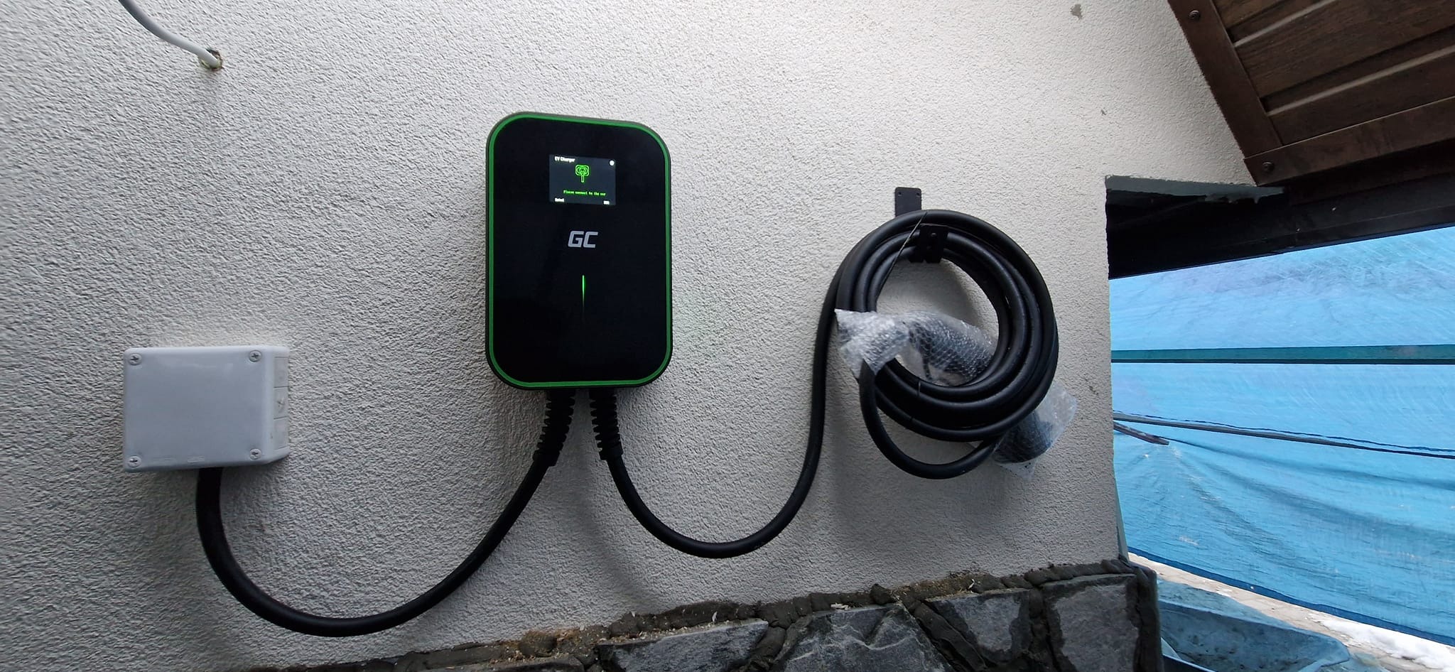 Zamontowana na zewnątrz, na teksturowanej ścianie, stacja ładowania pojazdów elektrycznych marki GC, z podłączonym przewodem zwiniętym na uchwycie. Obok widoczna puszka elektryczna.