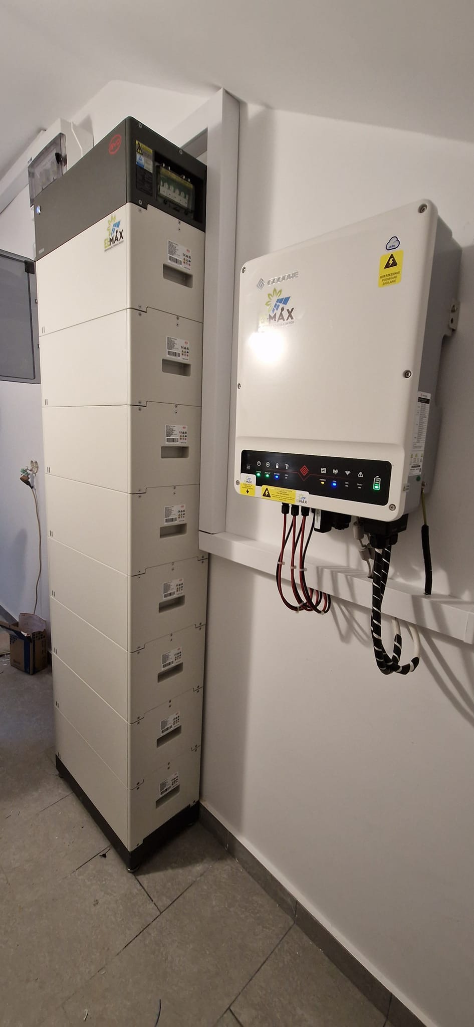 Biały modułowy magazyn energii E-MAX ułożony pionowo obok białego inwertera GoodWe na ścianie w pomieszczeniu.
