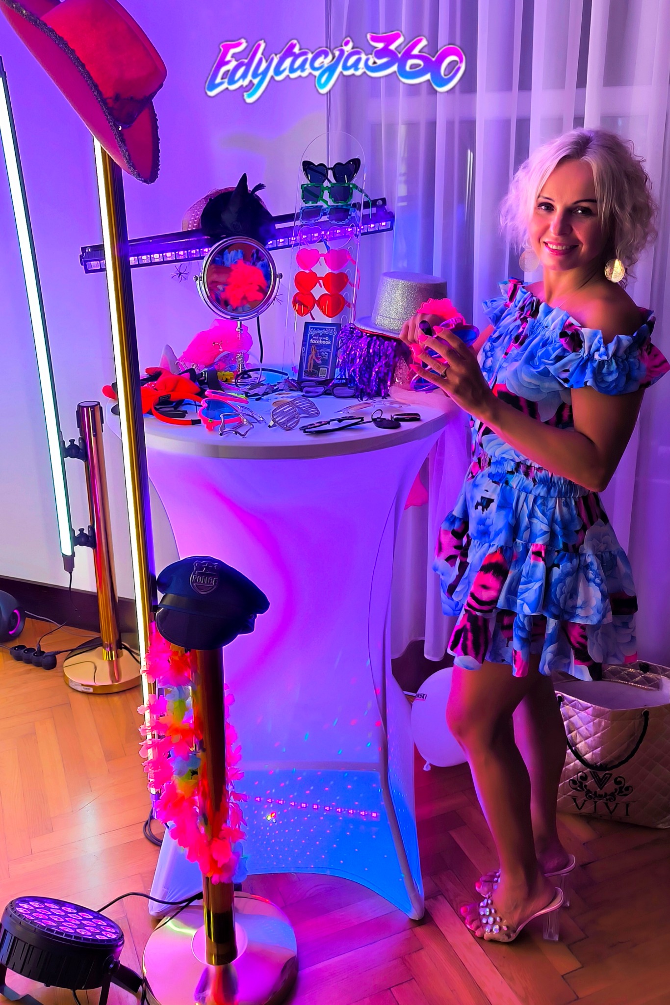 Uśmiechnięta blondynka w kwiecistej sukience stoi obok stołu z rekwizytami: kapelusze, okulary, lusterko, oświetlonego neonowym światłem, obok stoi statyw z czapką policyjną i girlandą kwiatów.