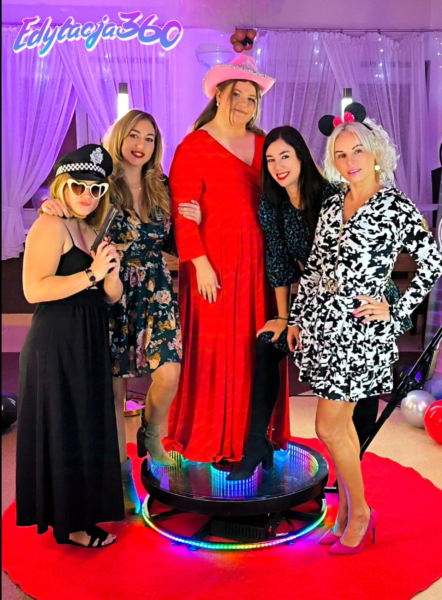 Pięć uśmiechniętych kobiet w kostiumach pozuje na obrotowej platformie z oświetleniem LED na czerwonym dywanie, w tle białe zasłony i balony.