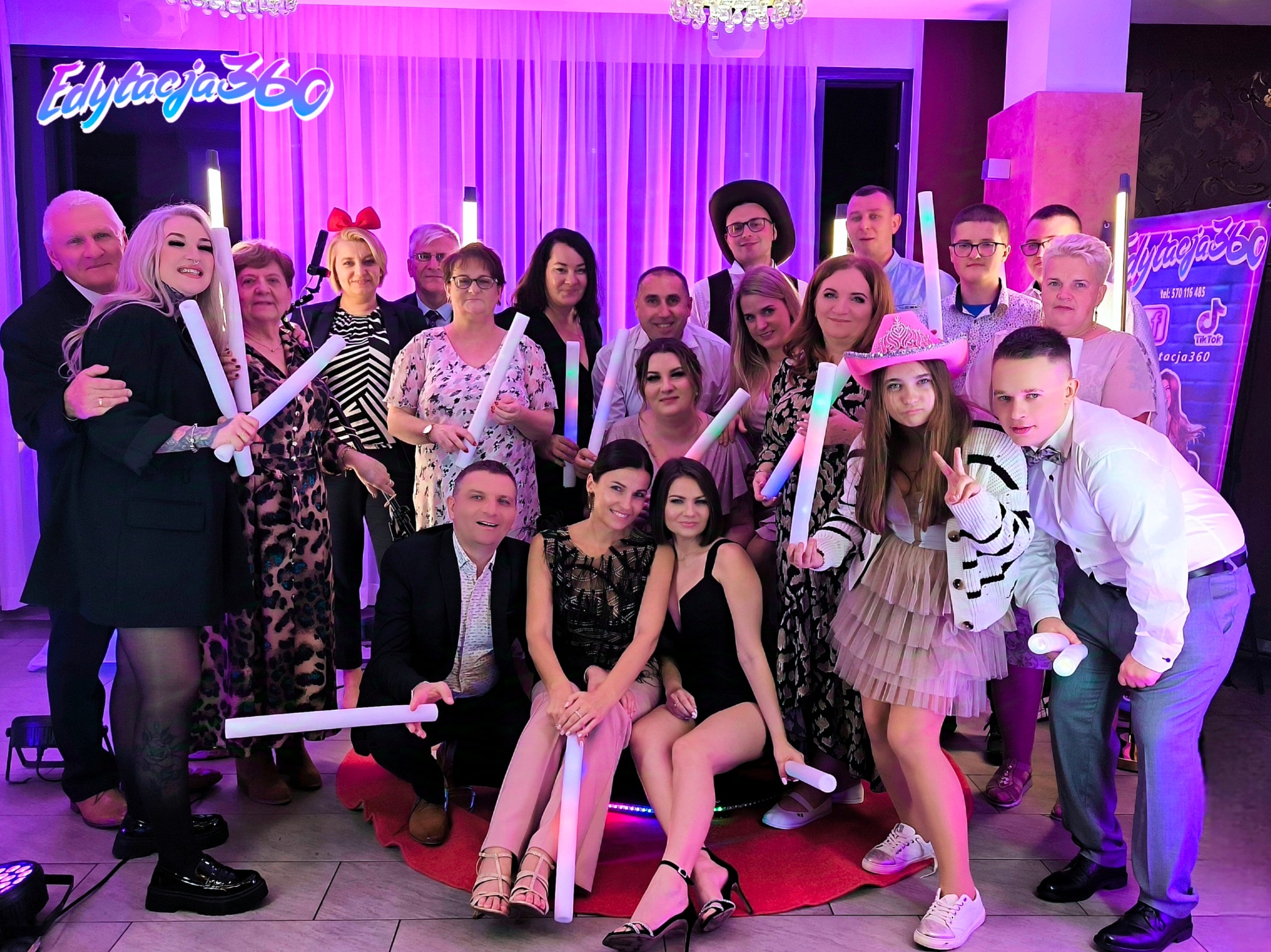 Grupowe zdjęcie uczestników imprezy z fotobudką, trzymających świecące pałeczki, na tle fioletowych zasłon i baneru reklamowego fotobudki Edytacja360.