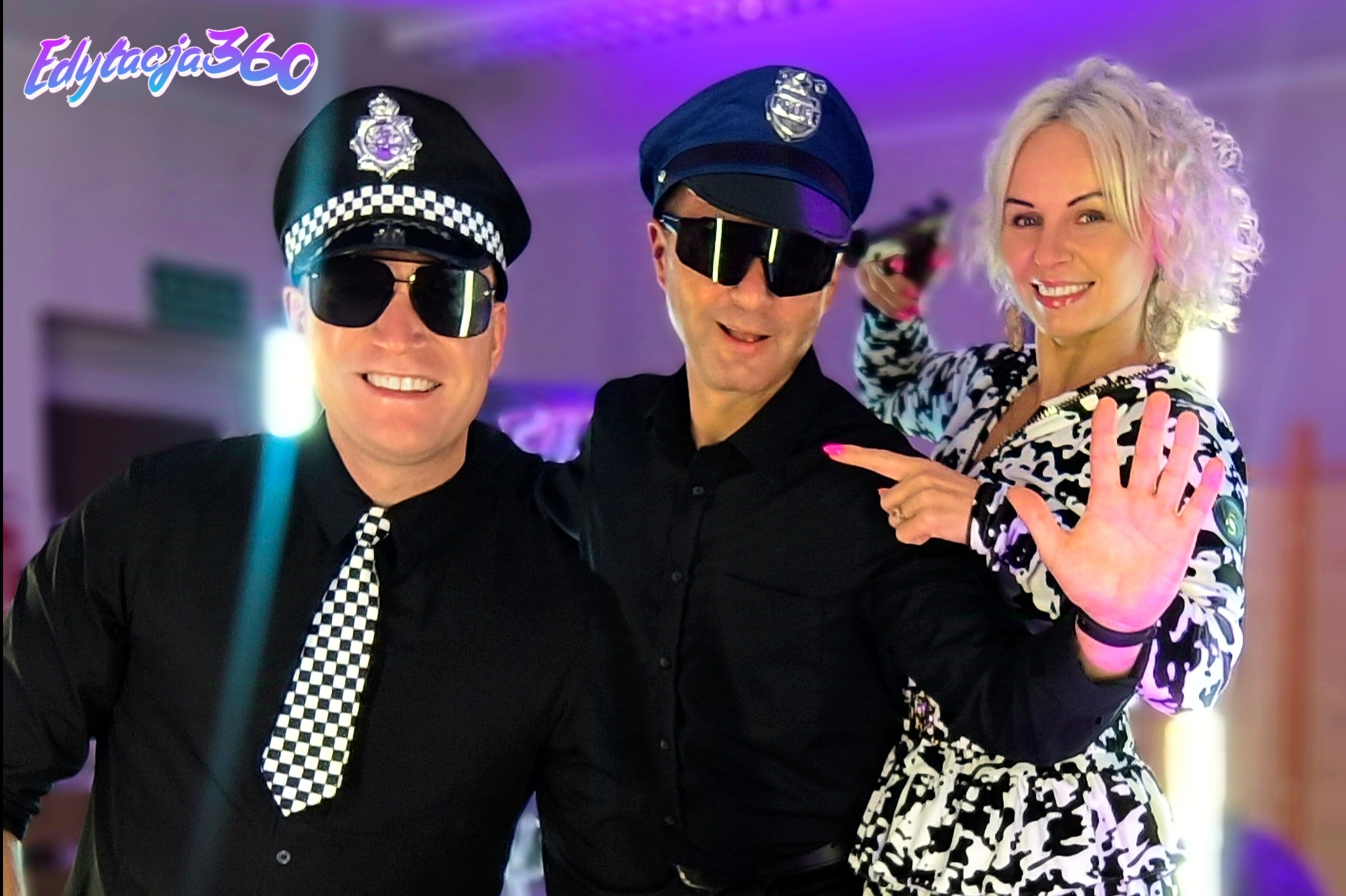 Trzy uśmiechnięte osoby pozują w przebraniach: dwóch mężczyzn w czarnych koszulach i czapkach policyjnych oraz kobieta w czarno-białej bluzce, z uniesioną dłonią i zabawką w kształcie pistoletu...