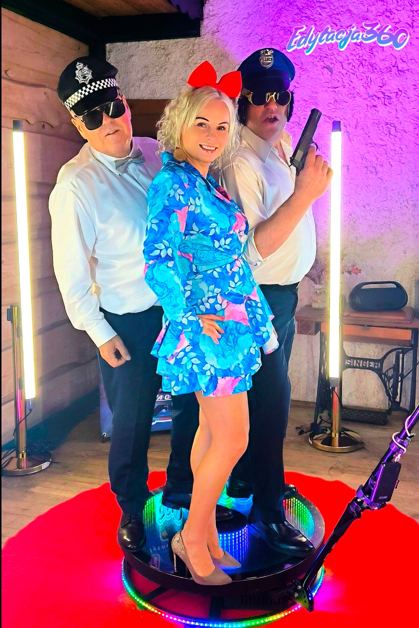 Trzy osoby pozują na obrotowej platformie z oświetleniem LED: kobieta z czerwoną kokardą we włosach i dwaj mężczyźni przebrani za policjantów, jeden z atrapą pistoletu, w tle stara maszyna...