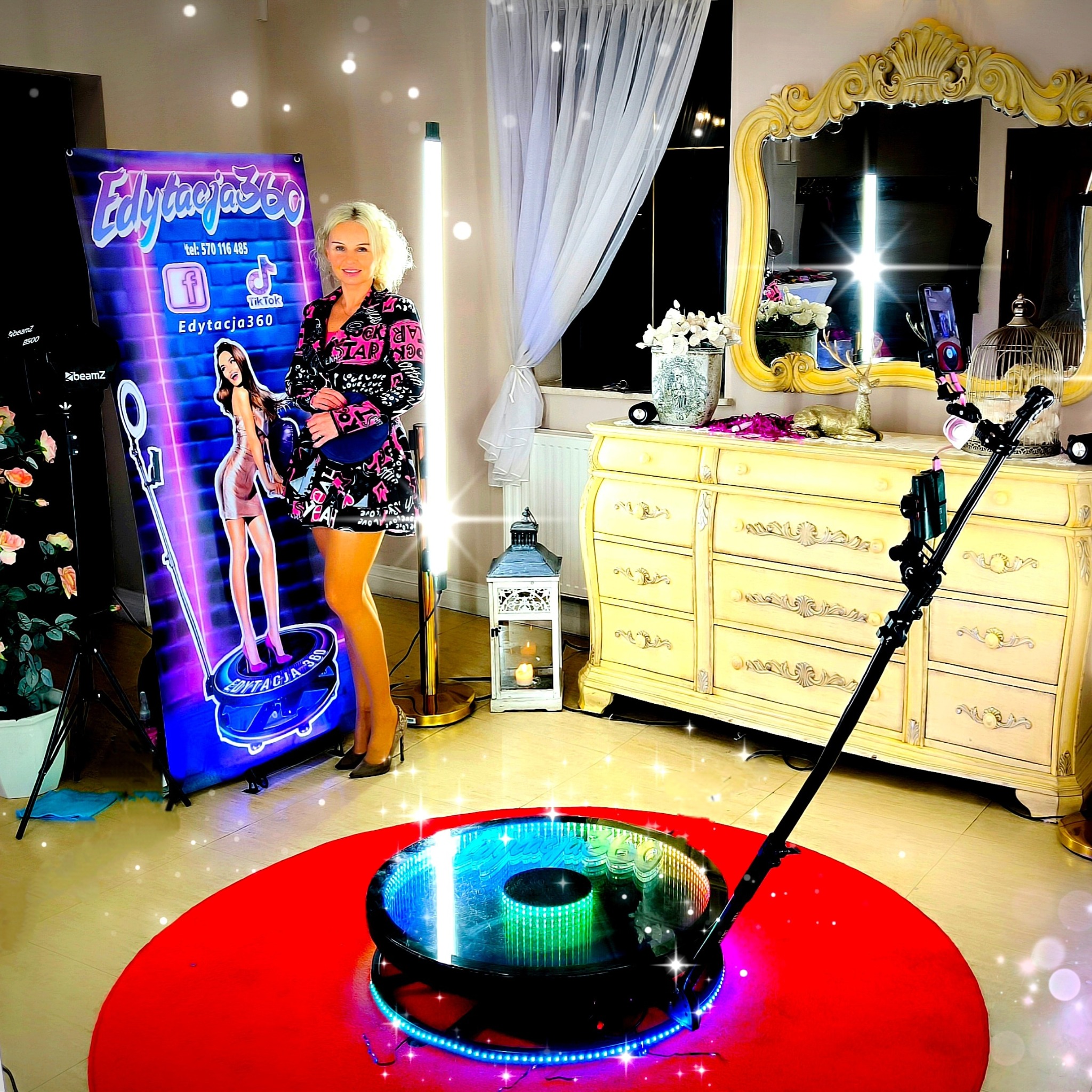 Kobieta pozuje obok podświetlanego baneru reklamowego z grafiką i nazwą 'Edytacja360', obok urządzenia do nagrywania filmów 360 na czerwonym dywanie, w tle komoda z lustrem i dekoracjami.