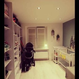 Artfinisz Daniel Szczyrba - Pokój dziecięcy z białymi meblami: łóżeczko, przewijak, regał, wózek, jasna podłoga i punktowe oświetlenie sufitowe.