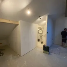 Poddasze w trakcie remontu: wygładzone ściany, widoczna konstrukcja drewniana, narzędzia i pracownik w tle.